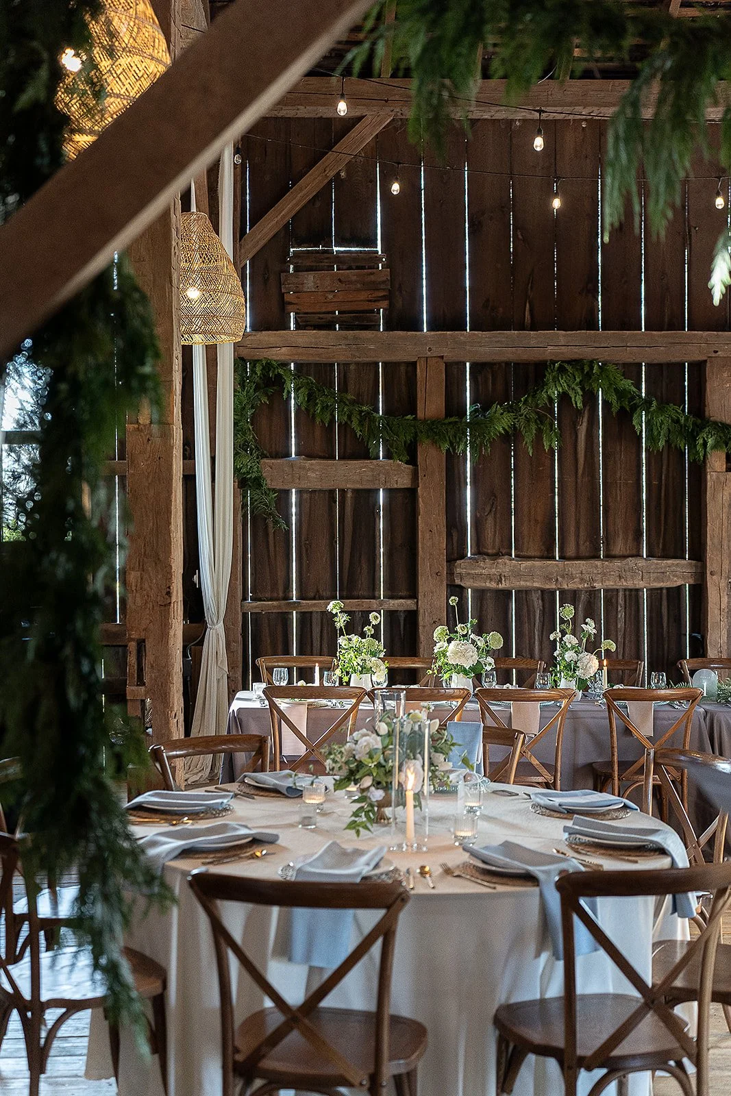 Indoor event space with round tables covered in white tablecloths, decorated with white and green floral centerpieces, set with white napkins, glasses, and gold utensils. The space has a rustic wooden interior with string lights hanging from the ceil