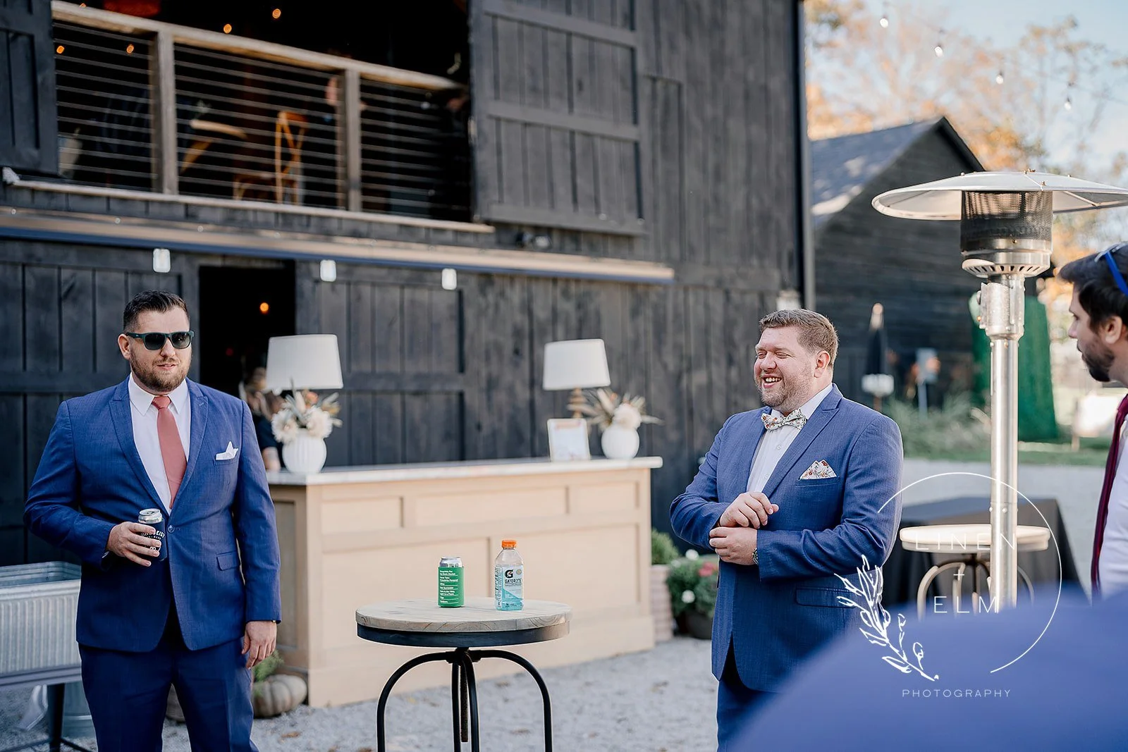 Three men in suits are having a conversation outdoors at a daytime social event, with a rustic wooden building and outdoor heater in the background.