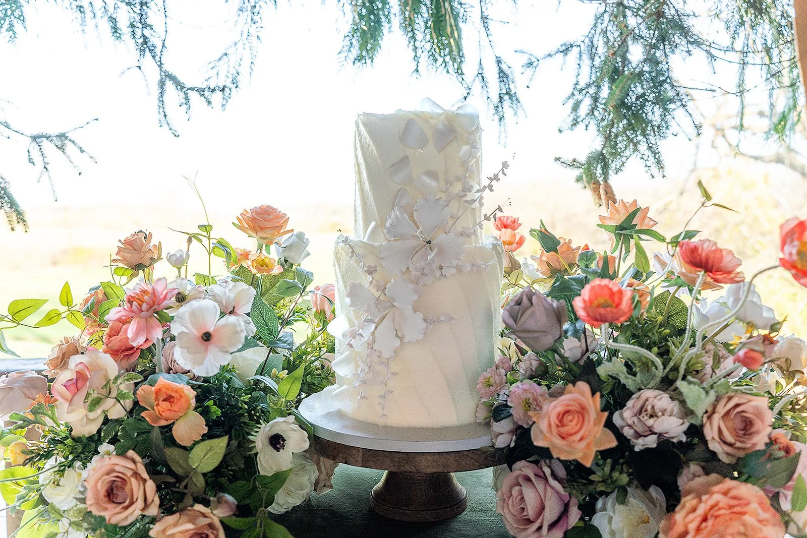 A white, two-tiered wedding cake decorated with white and pastel-colored sugar flowers, surrounded by a variety of pink, peach, white, and purple fresh flowers, with a grassy outdoor scene in the background.