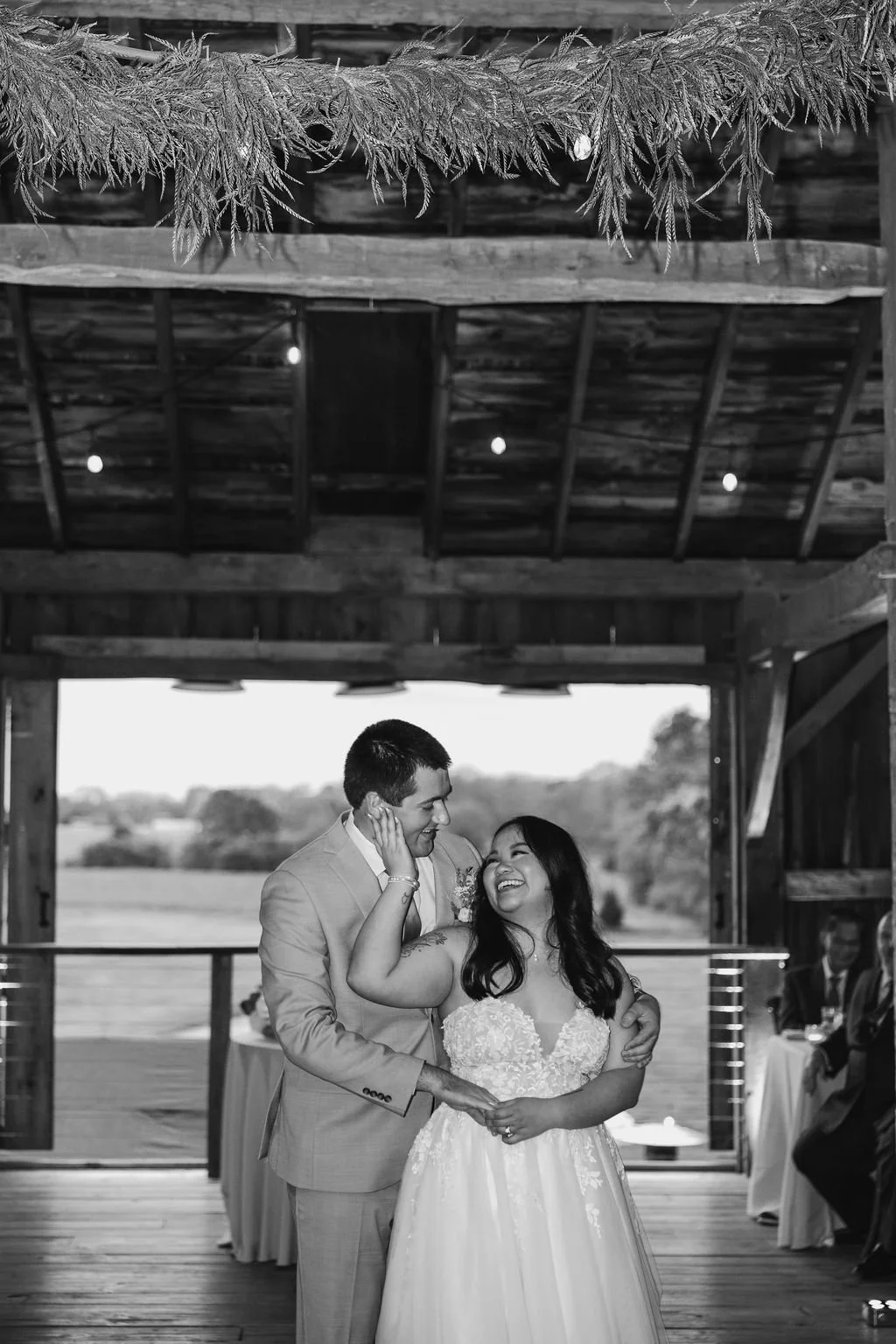 A black and white photo of a bride and groom dancing together inside a rustic wooden barn. The groom is wearing a suit and gently holding the bride's hand, while the bride, in a lace wedding dress, smiles joyfully at him at a 19th century estate