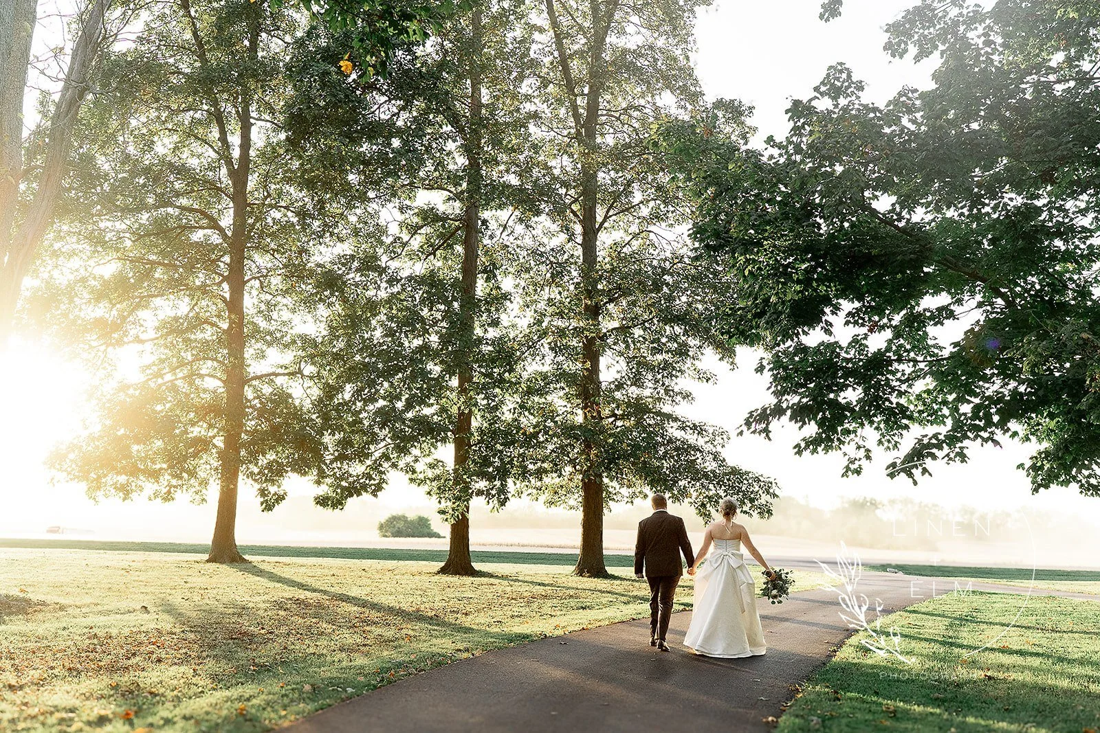 A bride and groom walking hand in hand along a park path during sunset, surrounded by tall trees and lush greenery.