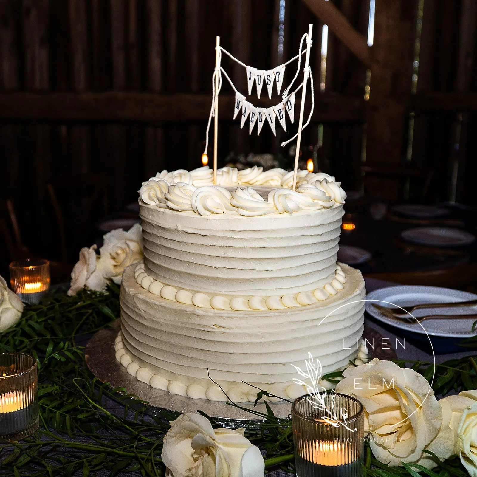 White wedding cake with piped frosting decoration, topped with a banner reading 'Just Married', surrounded by white roses and lit candles
