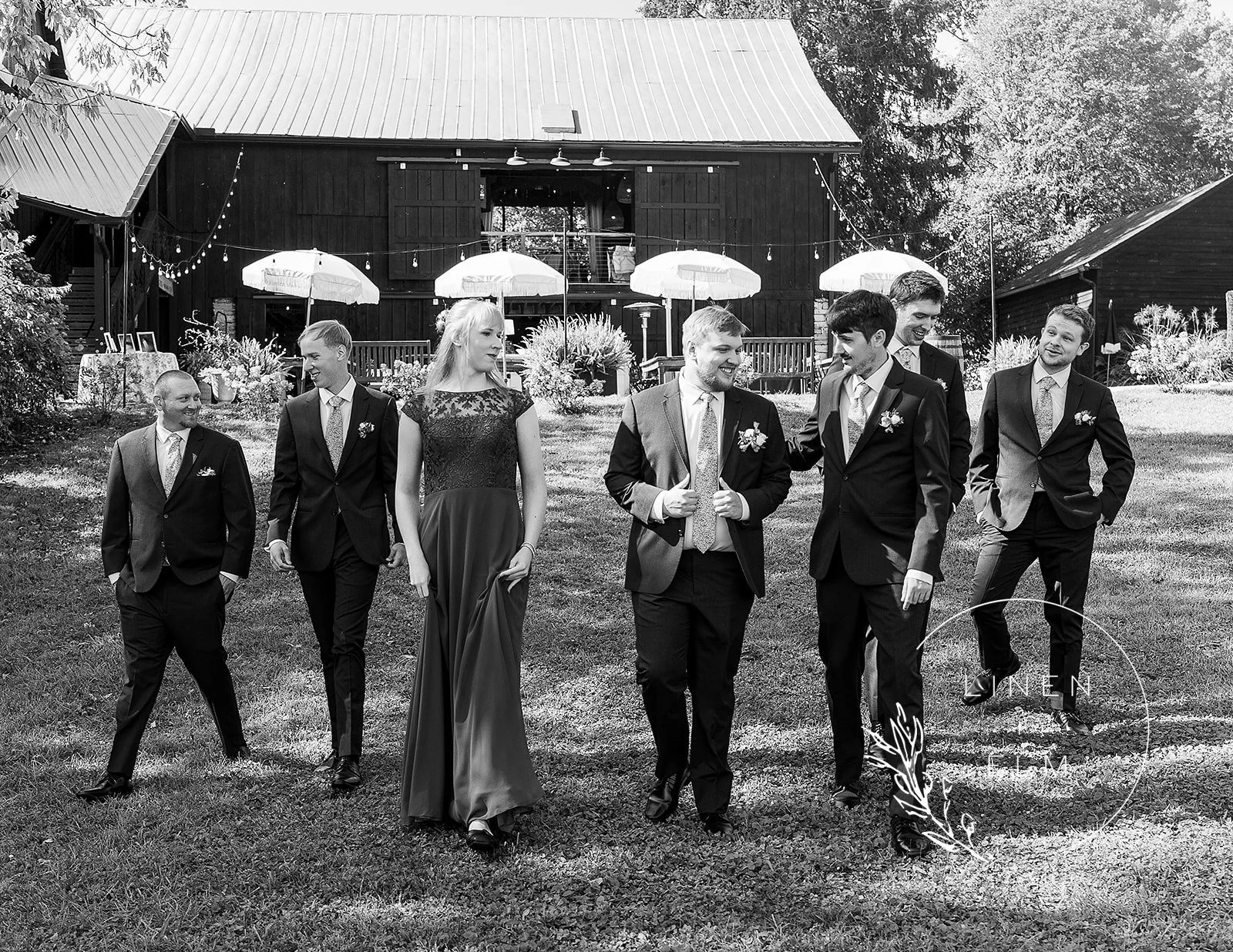 A black and white photo of a bride and six groomsmen walking together on a grassy outdoor area during a wedding celebration. The bride is wearing a long gown, and the groomsmen are in suits. The scene is decorated with umbrellas, string lights, and a