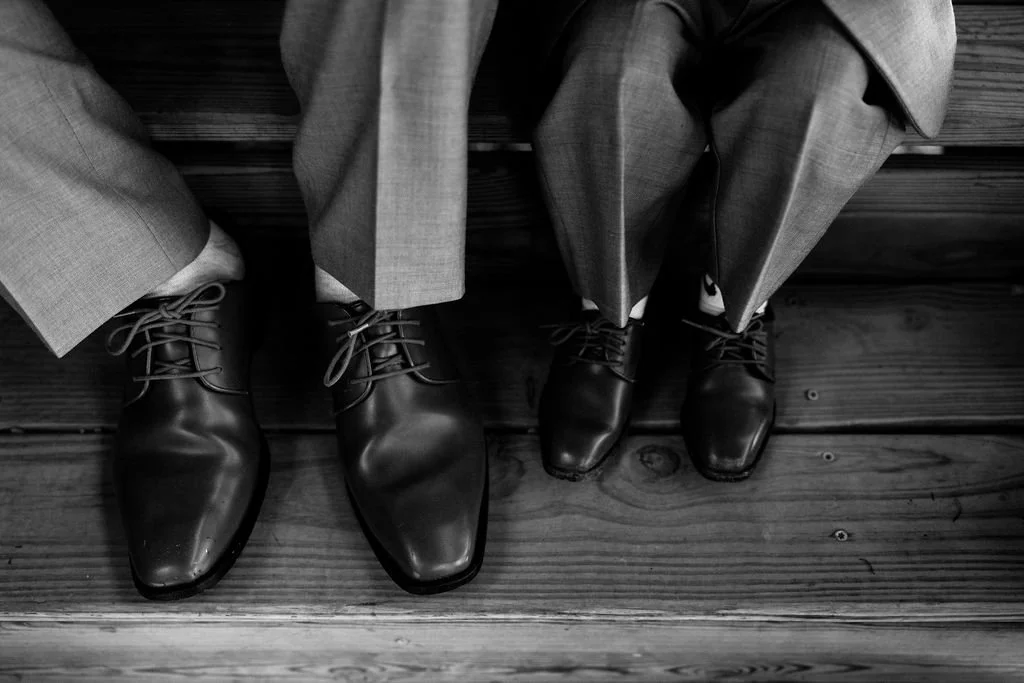 Two people sitting side by side, both wearing dress shoes and suit pants, with their feet visible on a wooden floor at a 19th century estate
