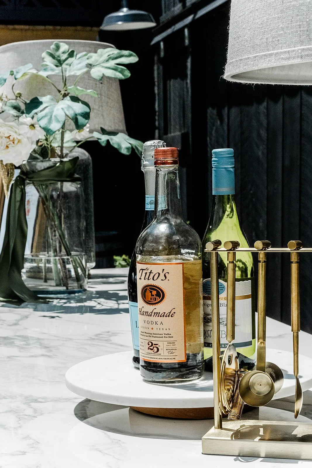A marble countertop with a vase of flowers, a bottle of Tito's handmade vodka, two wine bottles with blue caps, and a gold utensil holder with spoons and tongs, against a dark wall with a hanging lamp at a 19th century estate