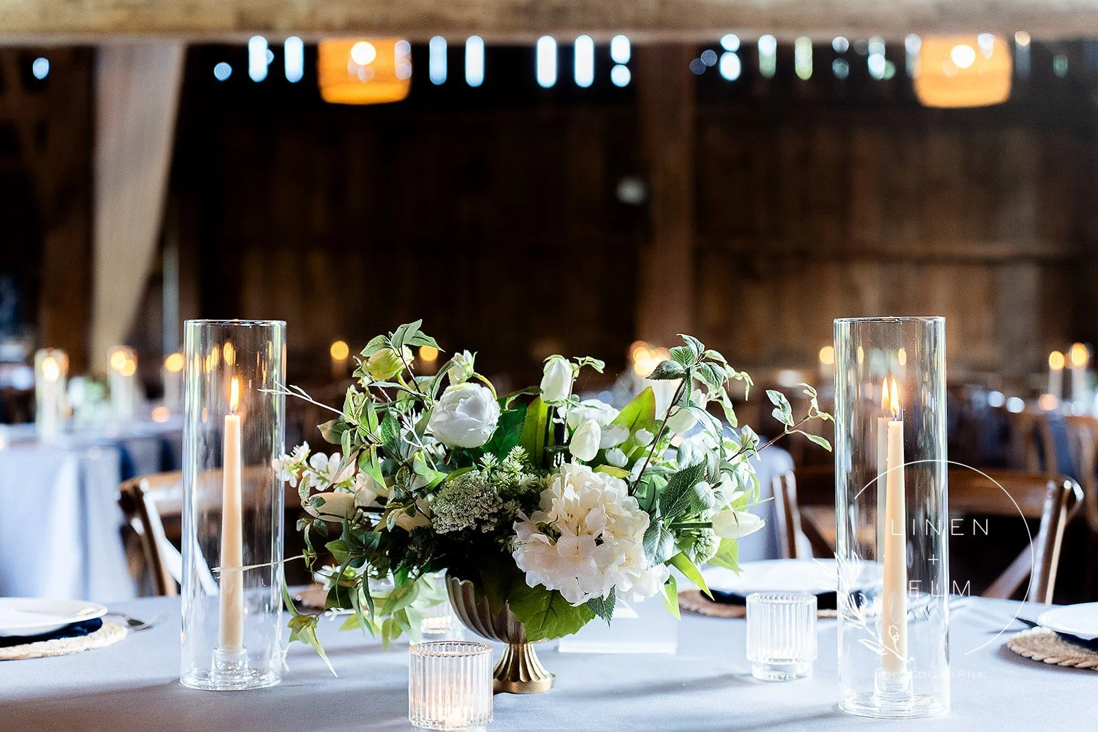Elegant table decorated with a floral centerpiece featuring white flowers and green foliage, surrounded by tall glass candleholders with lit candles, set in a rustic wooden venue with round tables and chairs in the background.