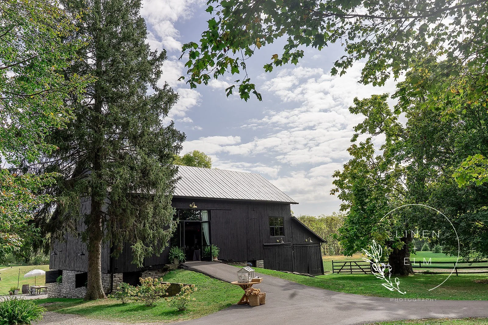 A black barn-style building with a metal roof situated among green trees on a cloudy day. The landscape includes a small table with a birdcage and a basket, a gravel driveway, and lush grass surrounding the building.