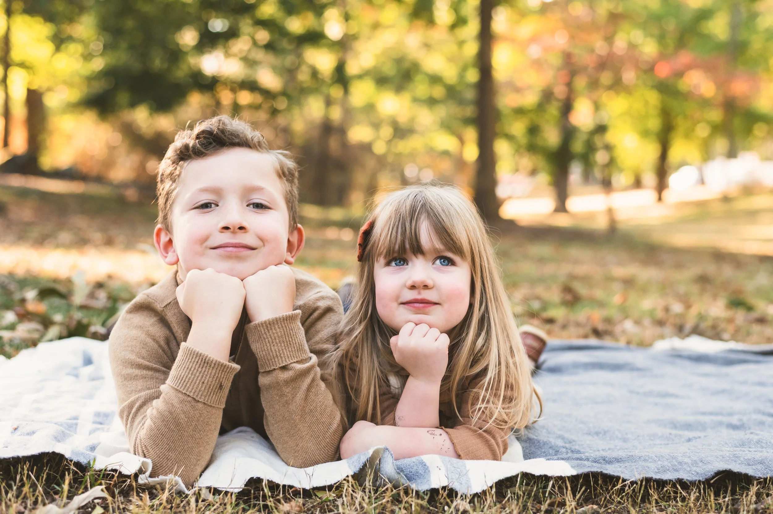 A boy and a girl lying on a blanket outdoors in a forested area during autumn, resting their chins on their hands and looking at the camera.