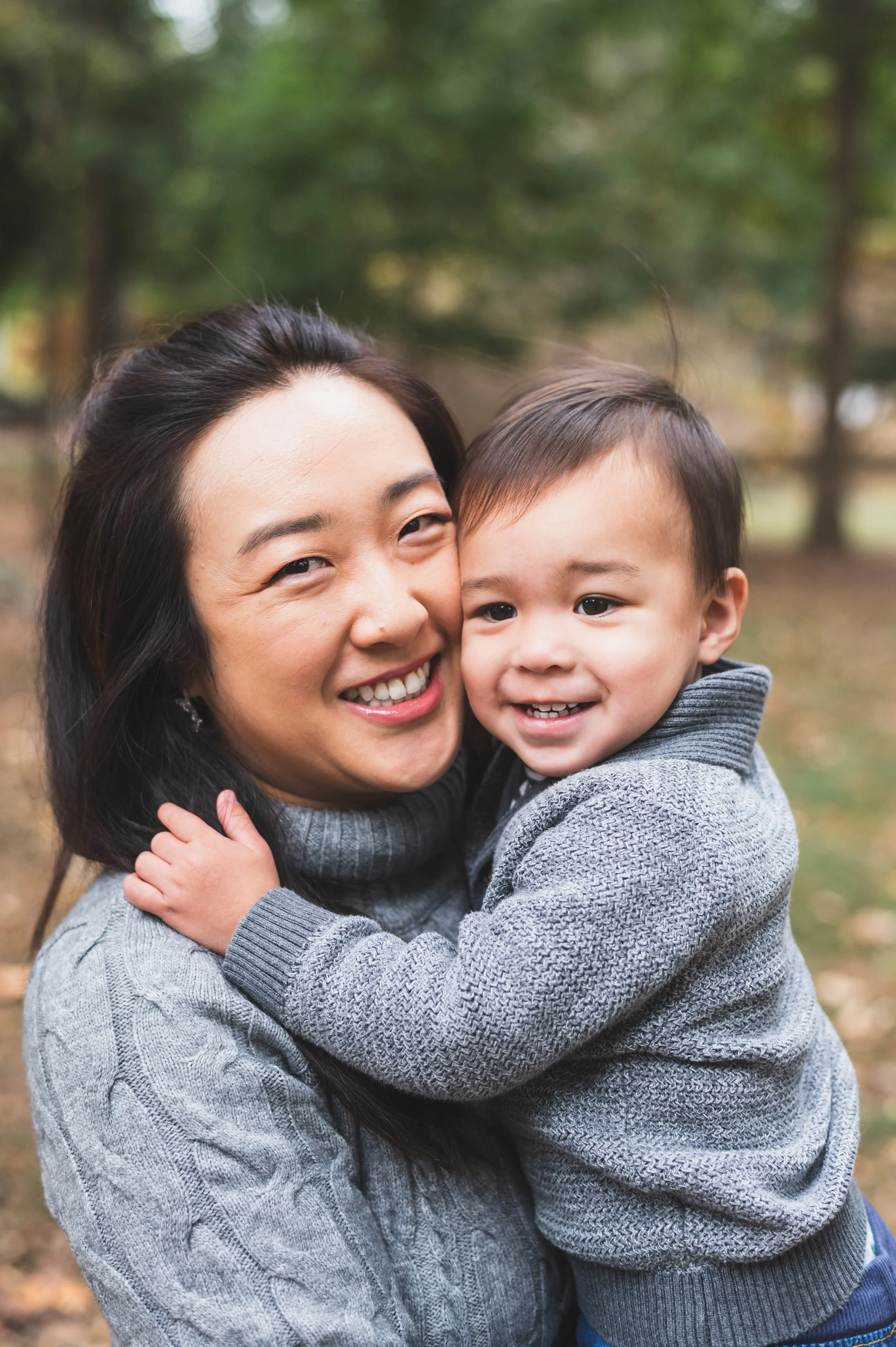 A woman and a young boy hugging outdoors in a wooded park, smiling and showing affection