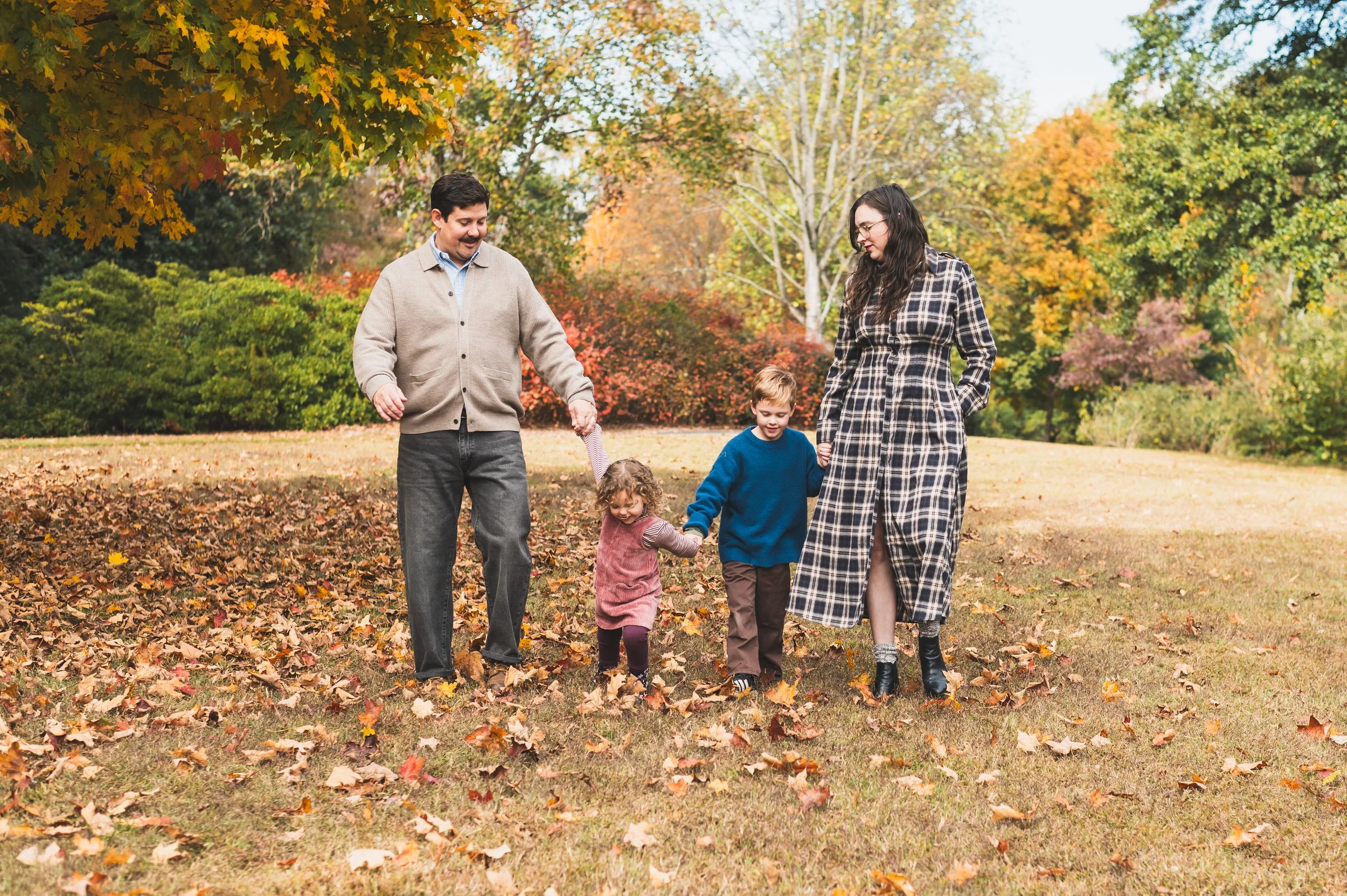 A family of four, including two children, walking in a park covered with fallen autumn leaves, surrounded by trees with colorful fall foliage.