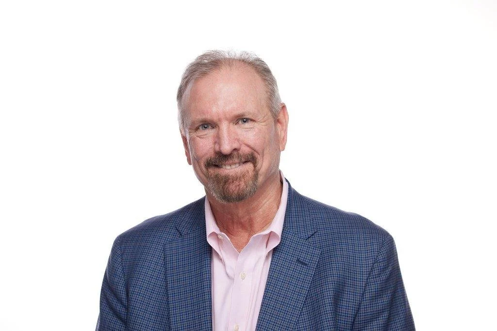 A middle-aged man with a beard and mustache smiling, wearing a blue checkered blazer and a light pink shirt, standing against a white background.