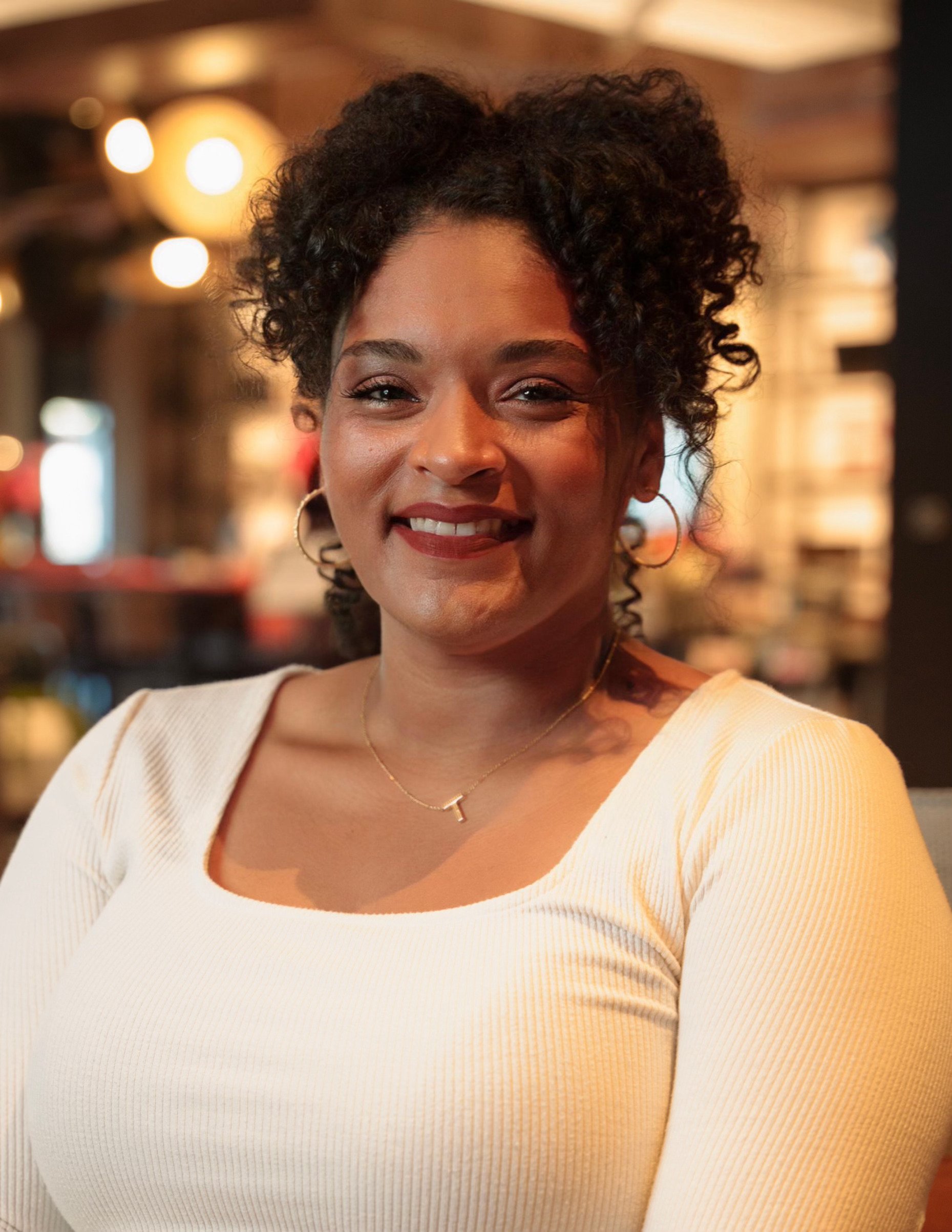 A woman with curly hair smiling at the camera in a cozy, warmly lit indoor space.