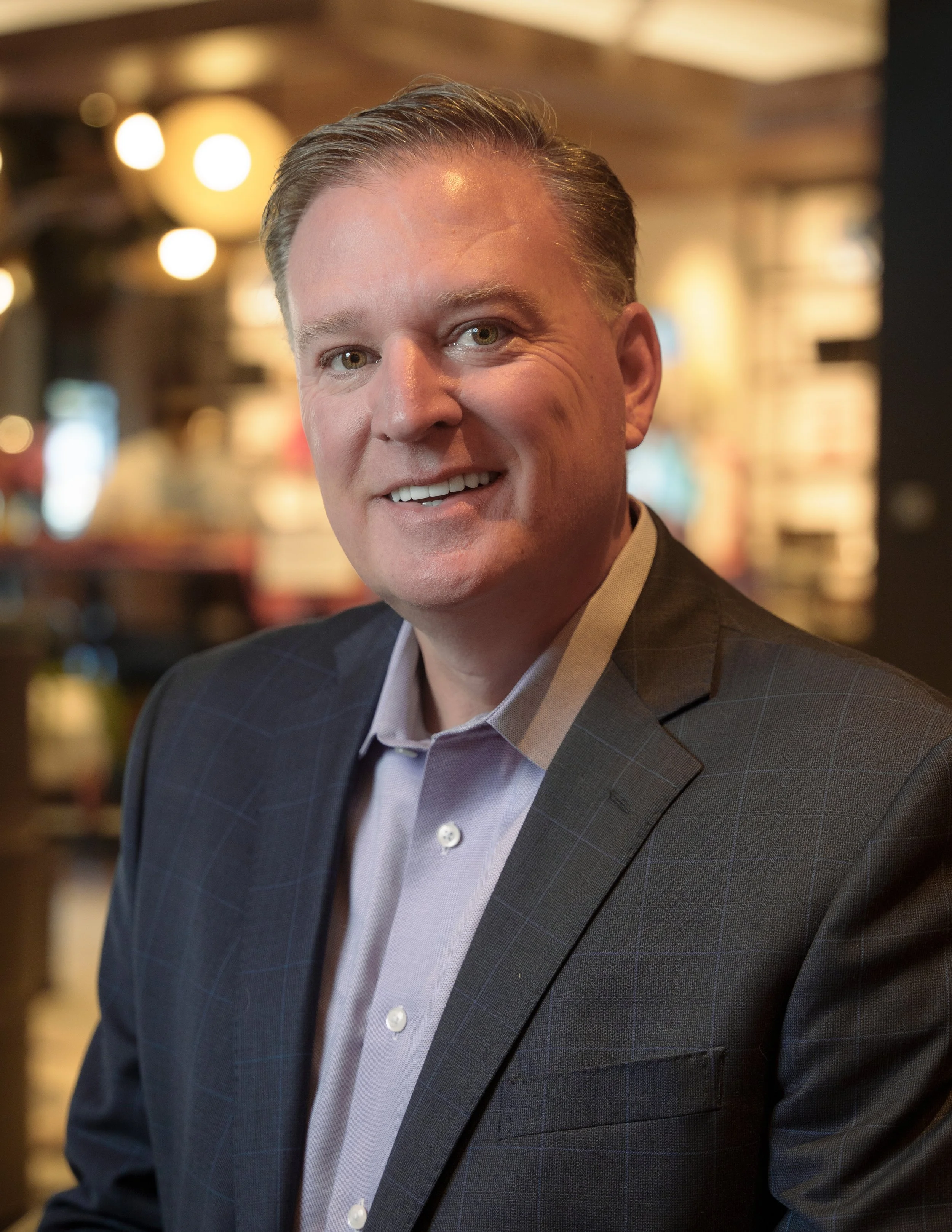 A man in a dark suit smiling in a warmly lit indoor setting.