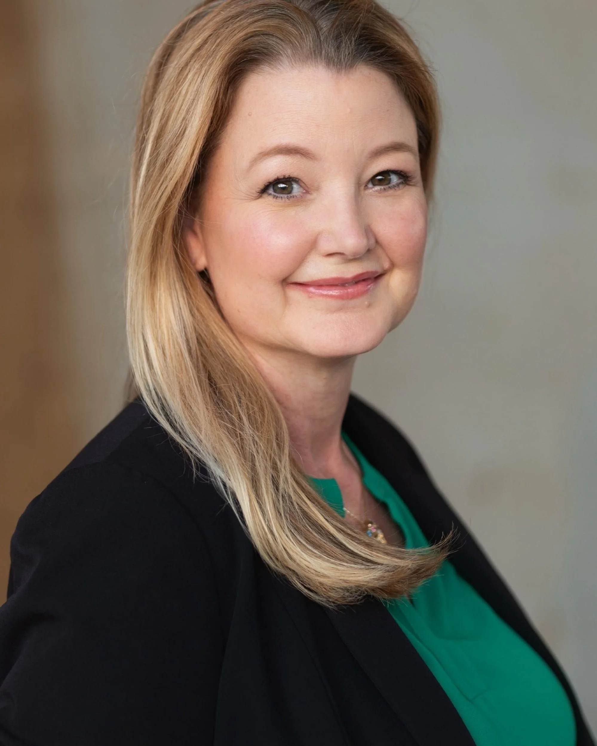 Close-up of a woman with shoulder-length blonde hair, wearing a black blazer and green top, smiling at the camera.