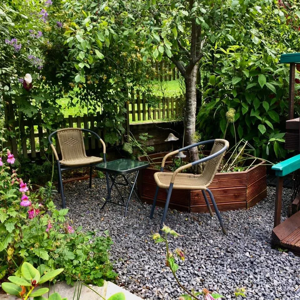 The Little India garden sitting area with two rattan chairs and a small glass table on gravel ground, surrounded by lush green plants and trees, with a wooden fence in the background.