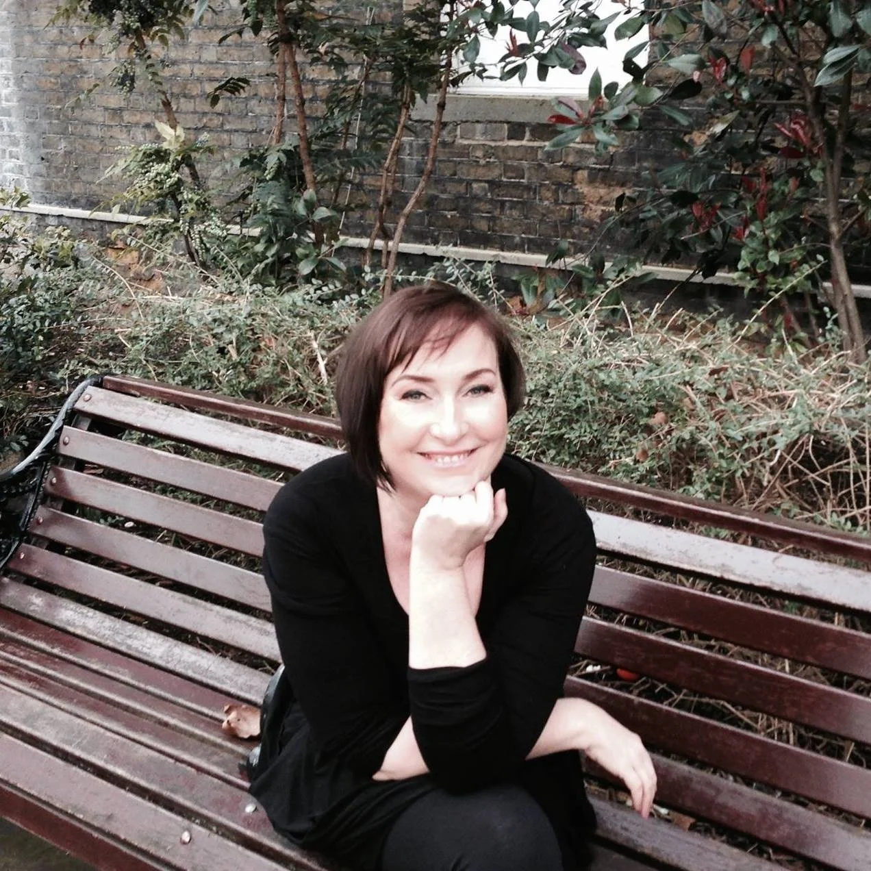 A woman with short brown hair and a black outfit sitting on a wooden park bench, smiling with her chin resting on her hand, in an outdoor garden setting with plants and a brick wall in the background.