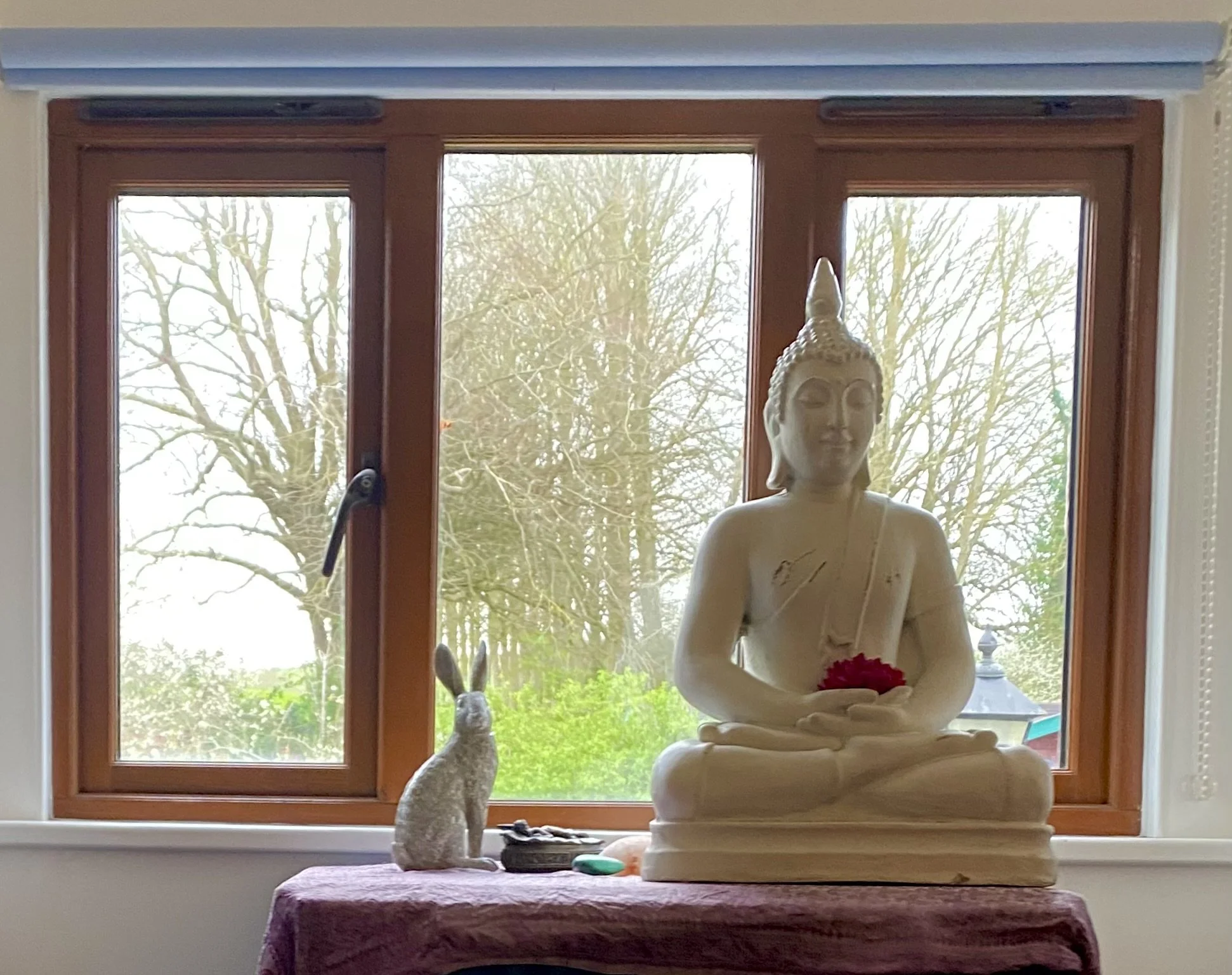 A Buddha Statue sitting in meditation pose, holding a flower with a hare statue next to it in front of a window overlooking the Dorset countryside at Spiritual Spa.
