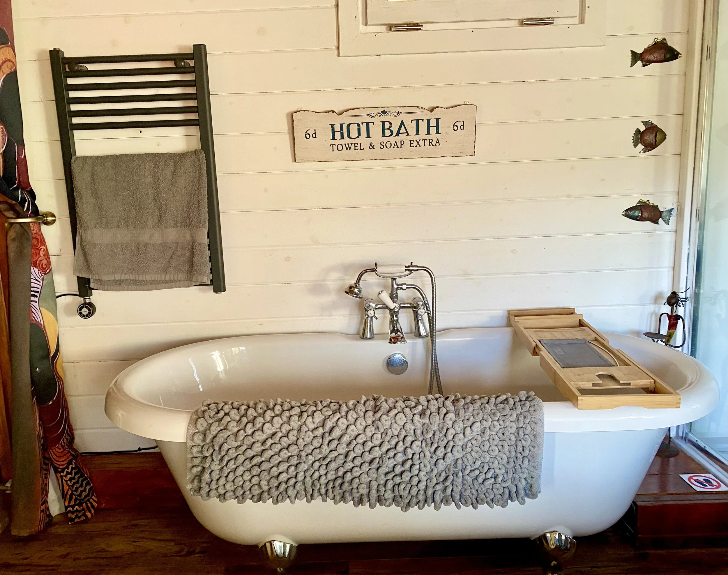 A vintage-style bathtub with curved edges, draped with a pebble-patterned towel, in a rustic bathroom. The wall behind is white wood paneling with a sign that reads 'Hot Bath Towel & Soap Extra' and three decorative fish figures. There's a silver faucet above the bathtub and a black towel radiator with a grey towel hanging on it. Small scattered items are visible nearby, including a wooden crate and some decorative elements.