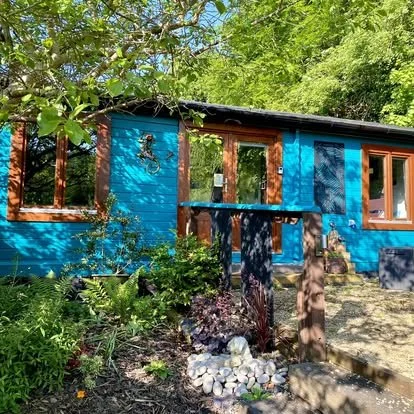 A blue wooden house with brown window frames in a lush garden with green trees and plants.