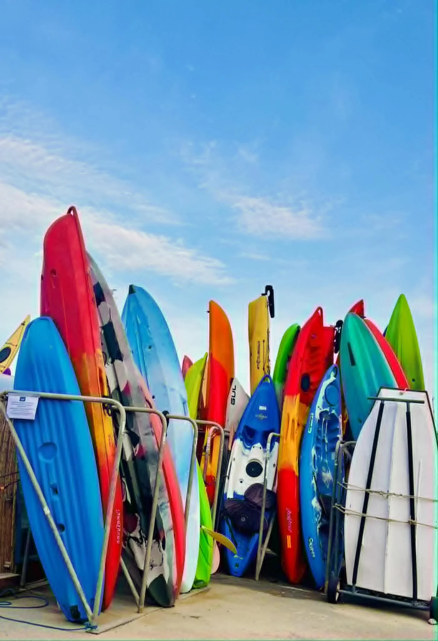 A collection of colorful surfboards standing upright outdoors, leaning against a metal rack, under a blue sky with some clouds.