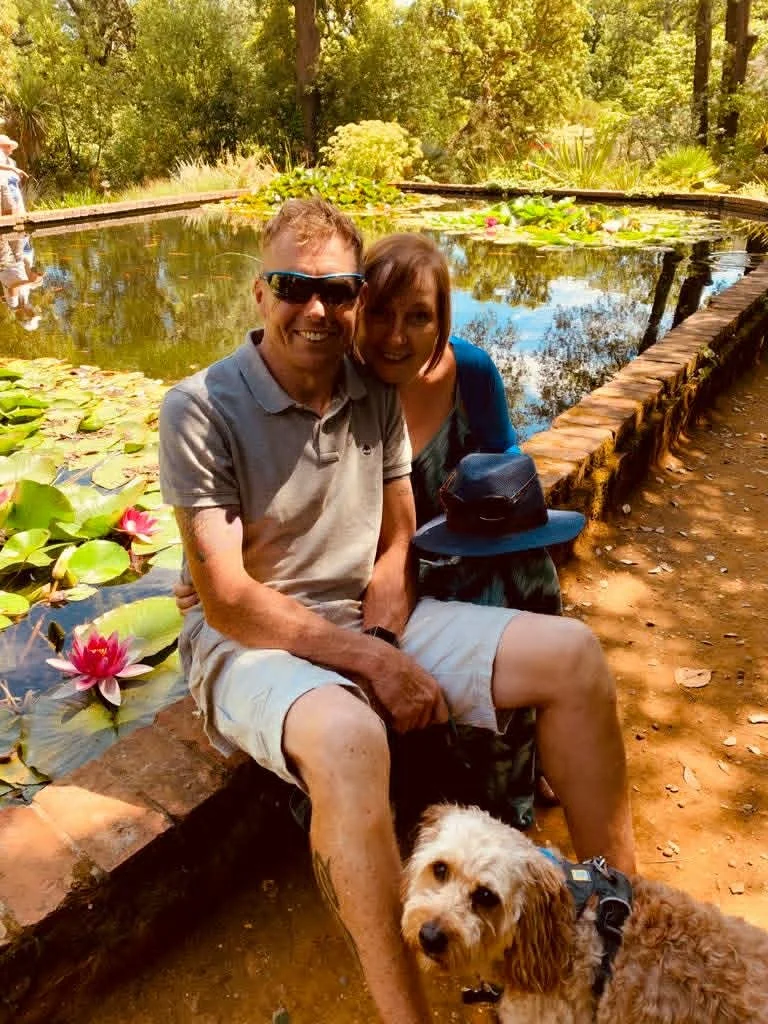 Nicky and her partner Mark sitting on a brick ledge next to a pond with lily pads and pink water lilies, outdoors with trees in the background. A dog is sitting in front of them, and a hat is placed on the woman's lap.