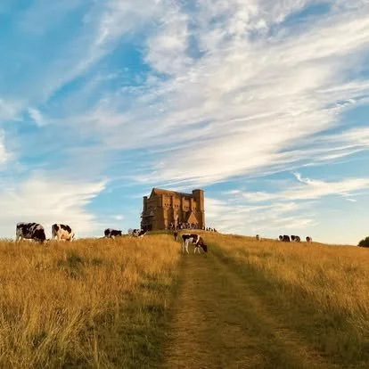 A castle on a hill with a dirt path leading up to it, surrounded by fields and grazing cows, under a partly cloudy sky.