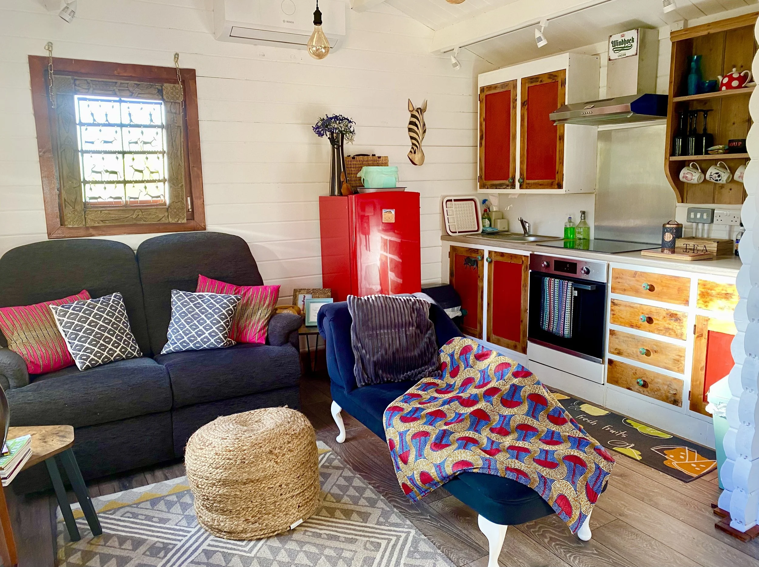 The living area and kitchen in the self-catering chalet Little Africa, there is a two-seater sofa, a chaise lounge and a small kitchen with fridge, stove, oven, sink and kettle