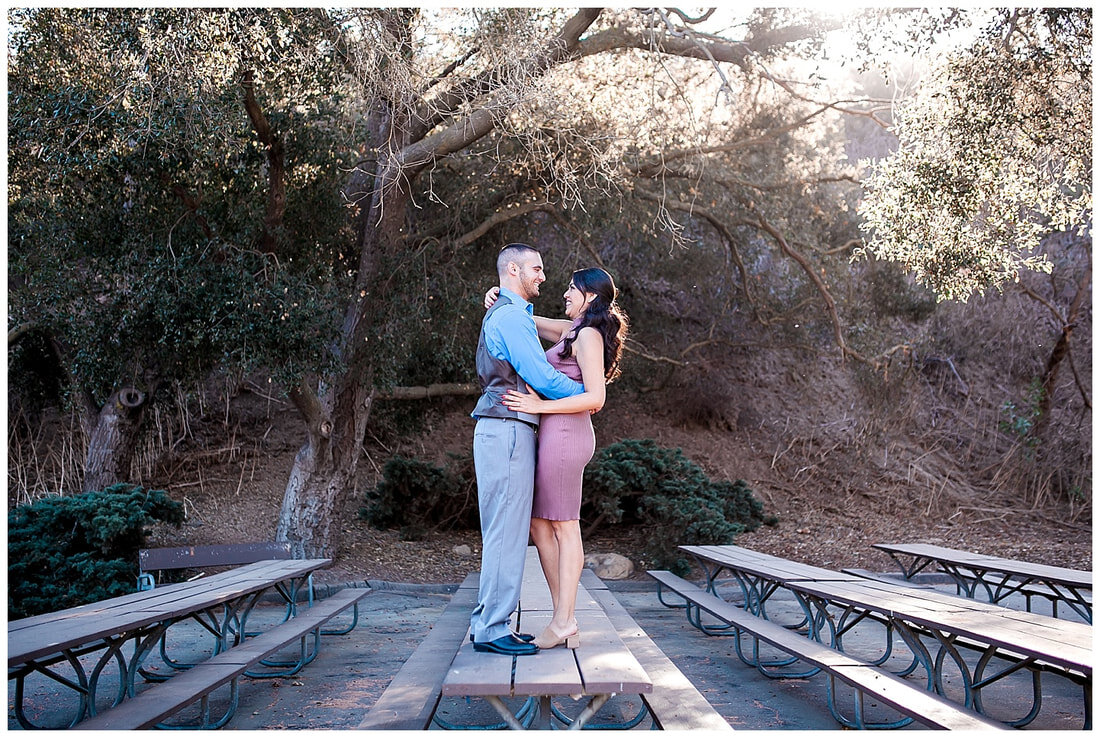  newly engaged couple smiling at one another as they stand atop a picnic table in the woods 