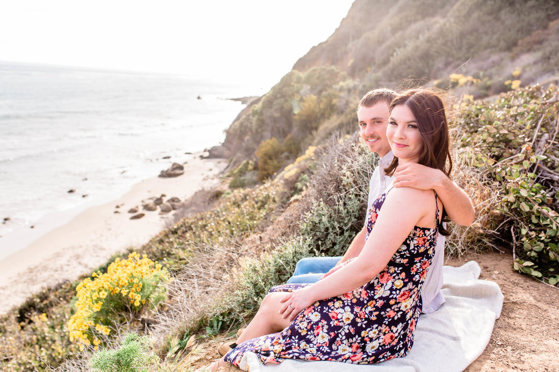  couple look to the camera with the ocean in the background and some yellow flowers 