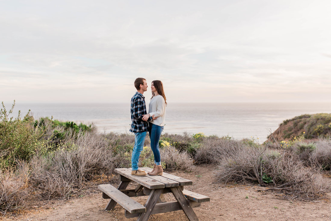  couple standing on top of a picnic table with the ocean behind them 