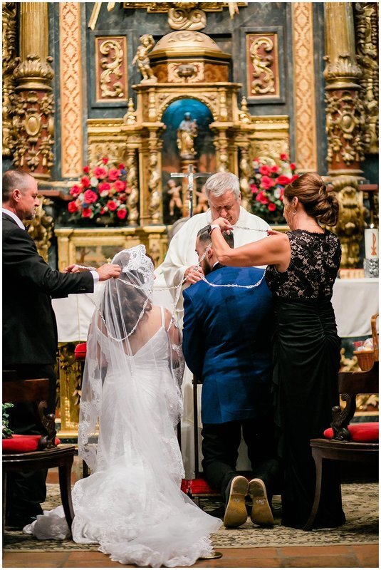  parents put the rosery tie on the bride and groom as they kneel before the priest 