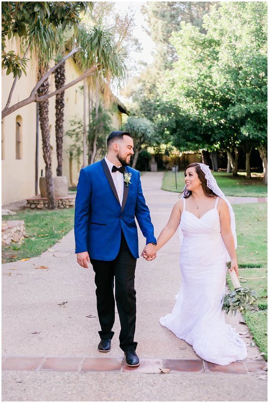  bride and groom walking down a path flanked by trees holding hands 