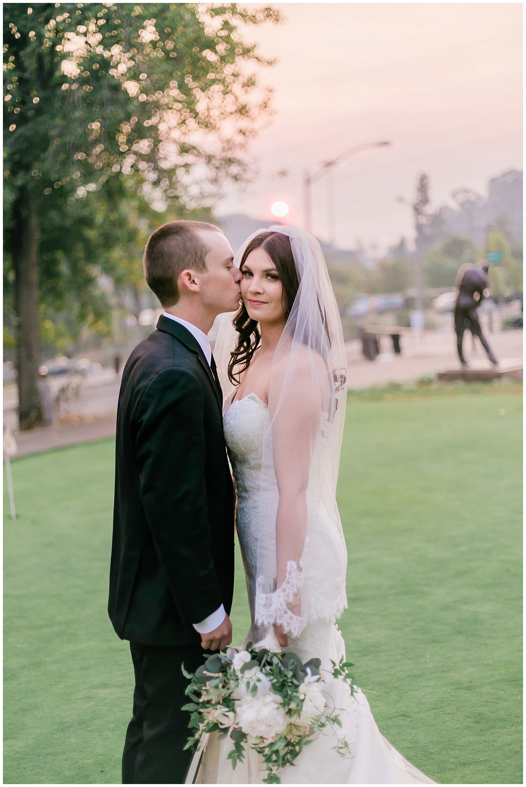  groom kisses his bride on the cheek while the sun sets behind them 