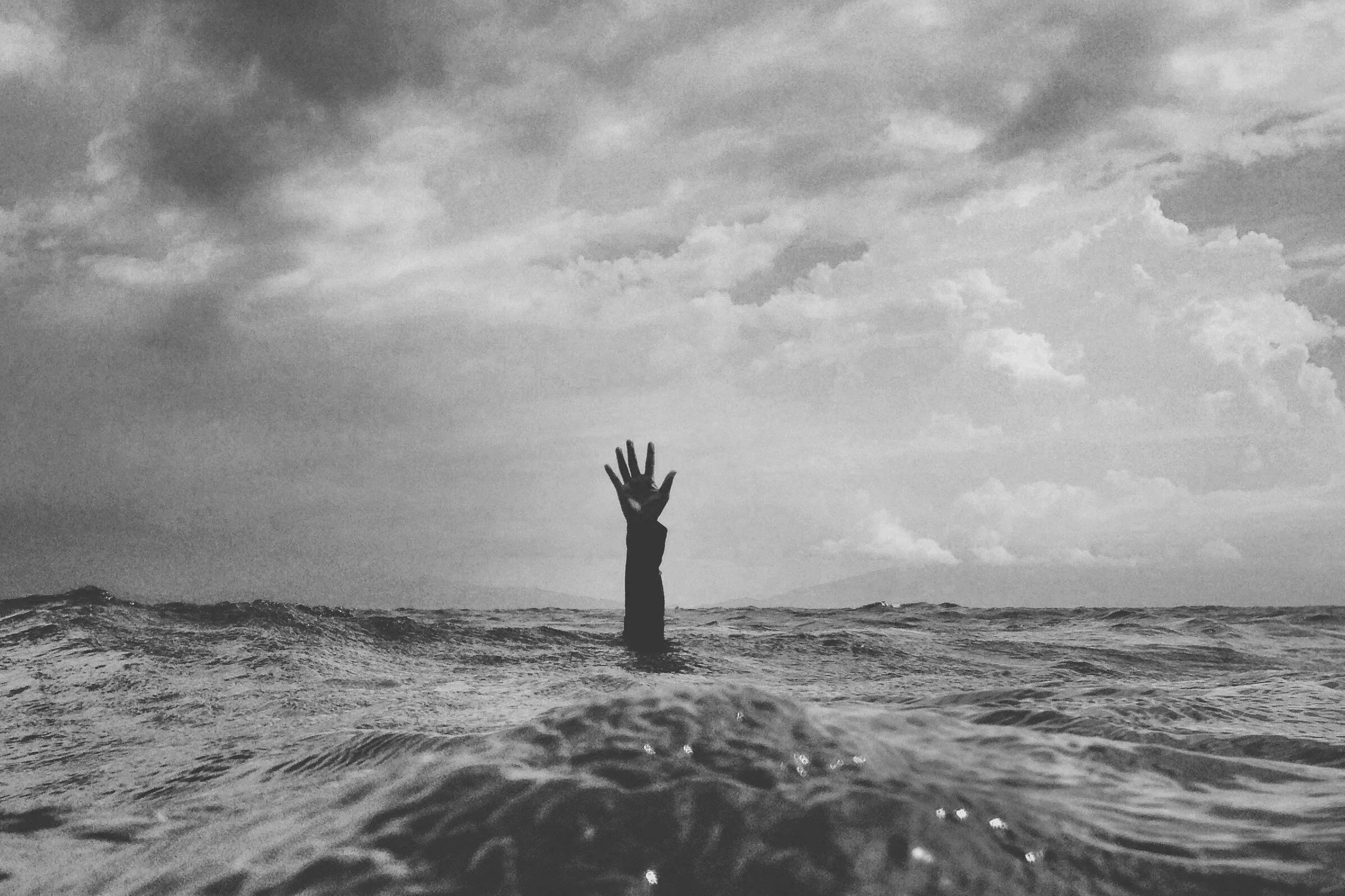A person is drowning in the ocean with a hand reaching out of the water, under a cloudy sky.