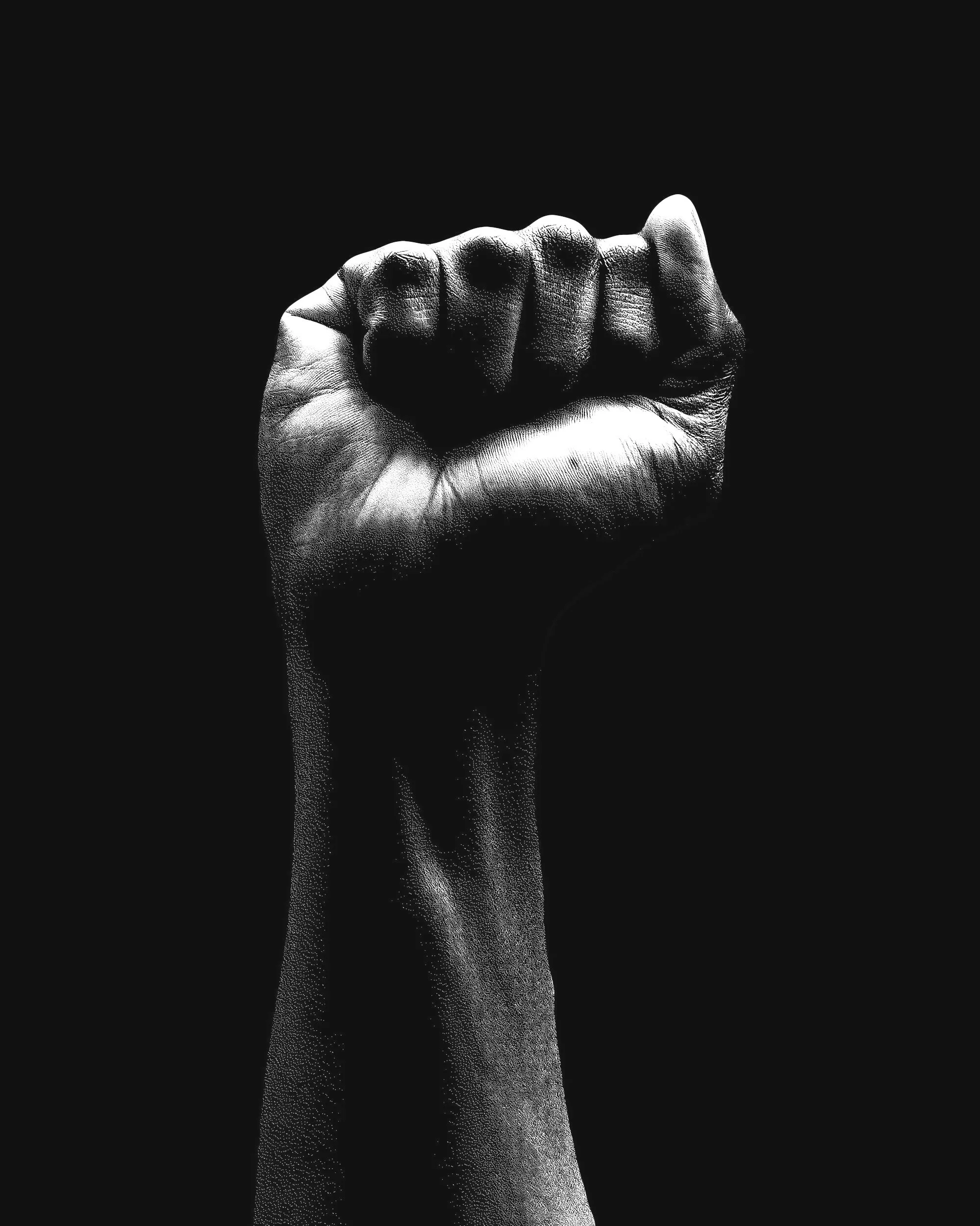 A black and white image of a human fist raised in a fist pump gesture against a dark background.