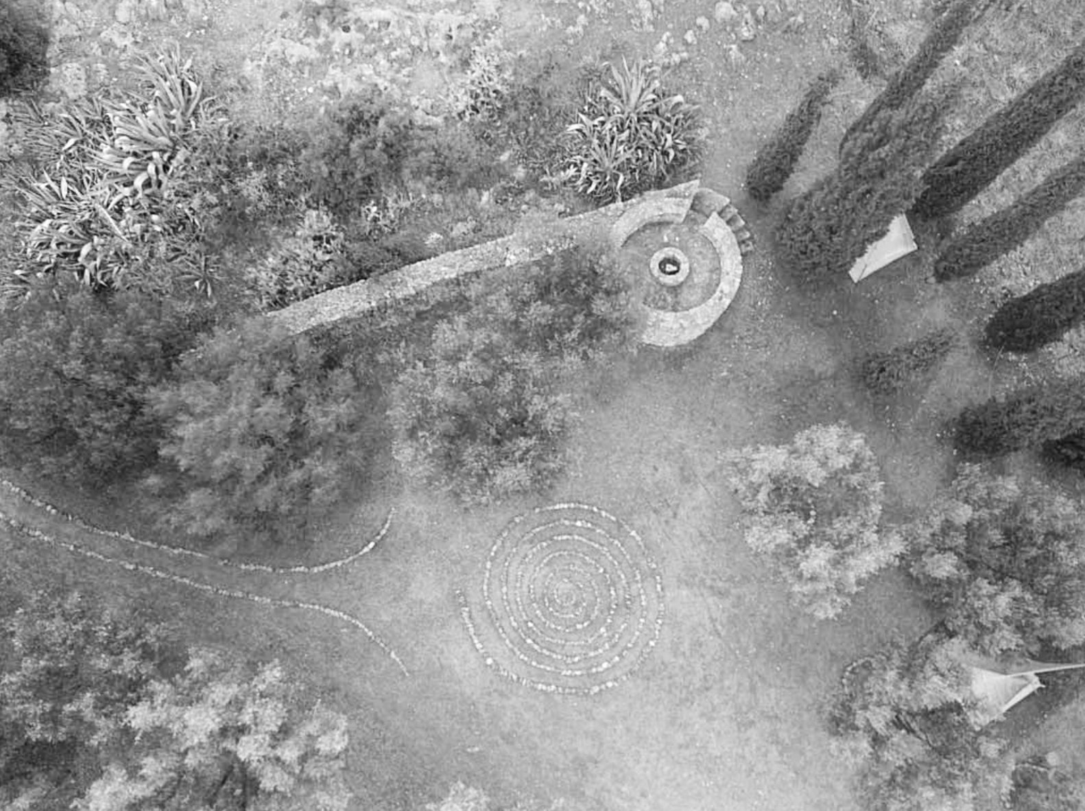 An aerial view of a landscaped outdoor area featuring spiral and semi-circular decorative patterns on the ground, surrounded by trees, bushes, and a stone spiral staircase.