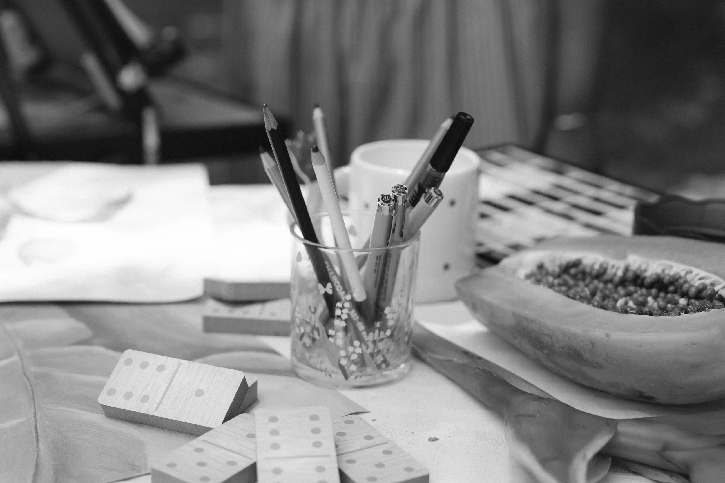 A desk with a glass holder filled with pens and pencils, some square tiles with dots, a domino, and a pie chart in a bowl, with a blurred background.