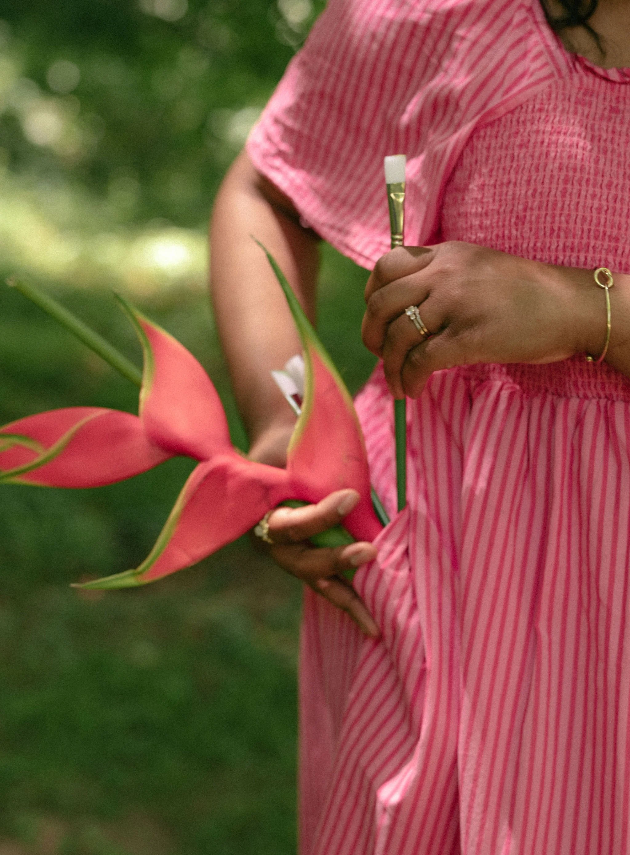 A woman in a pink and white striped dress holding a paintbrush near a pink flower, with green foliage in the background.