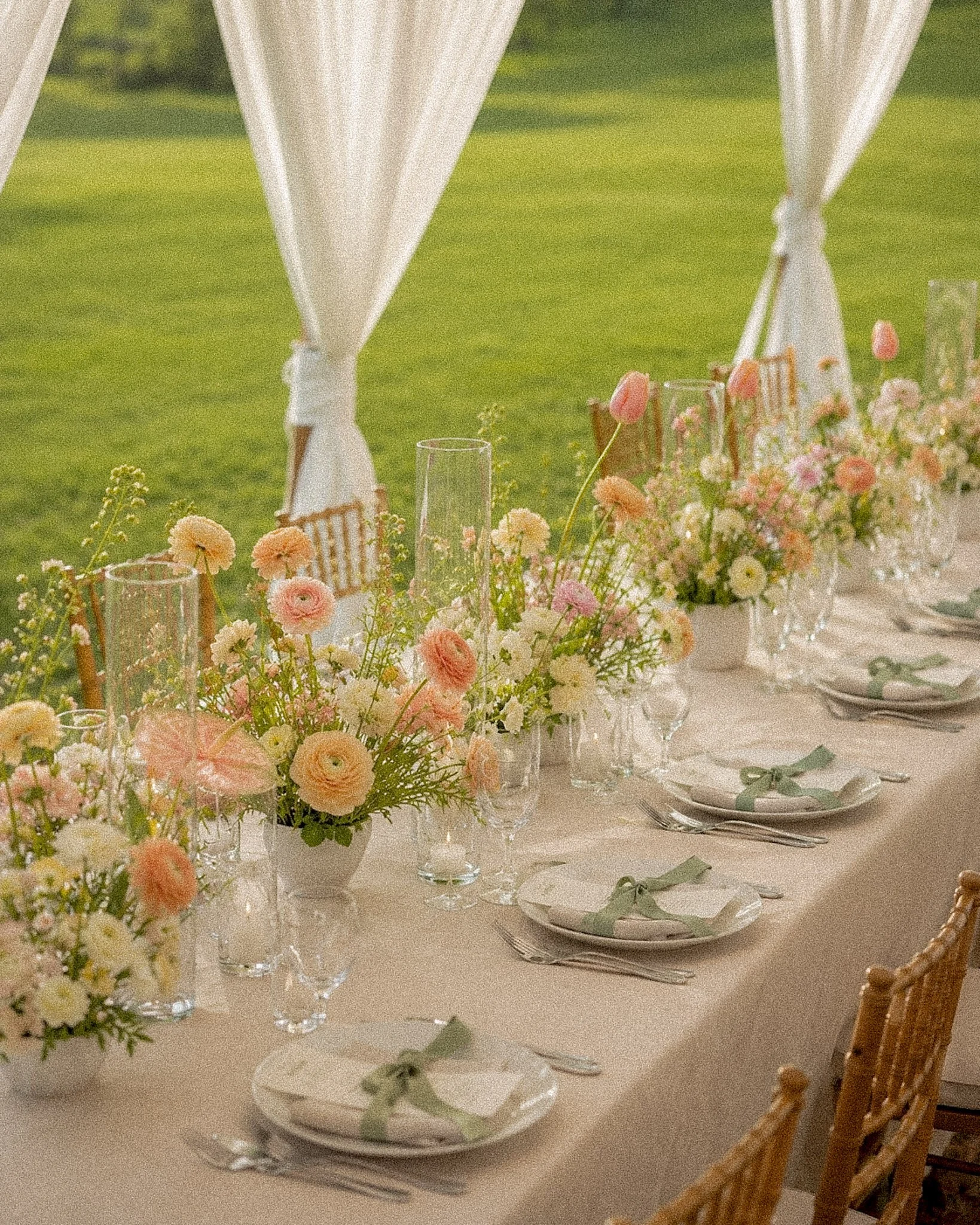 Close-up of a decorated dining table with floral arrangements, glassware, and plates, set outdoors against a grassy background for a wedding or celebration.