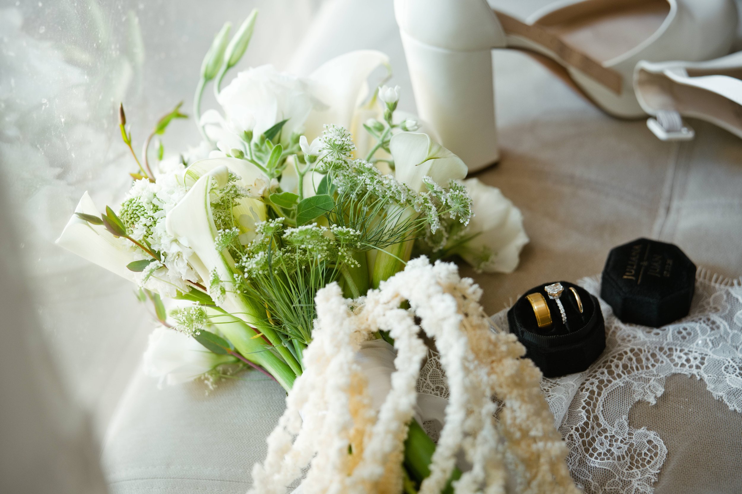 A floral arrangement with white flowers and green leaves on a lace tablecloth, near a white vase, a jewelry box, and a black lace ribbon.
