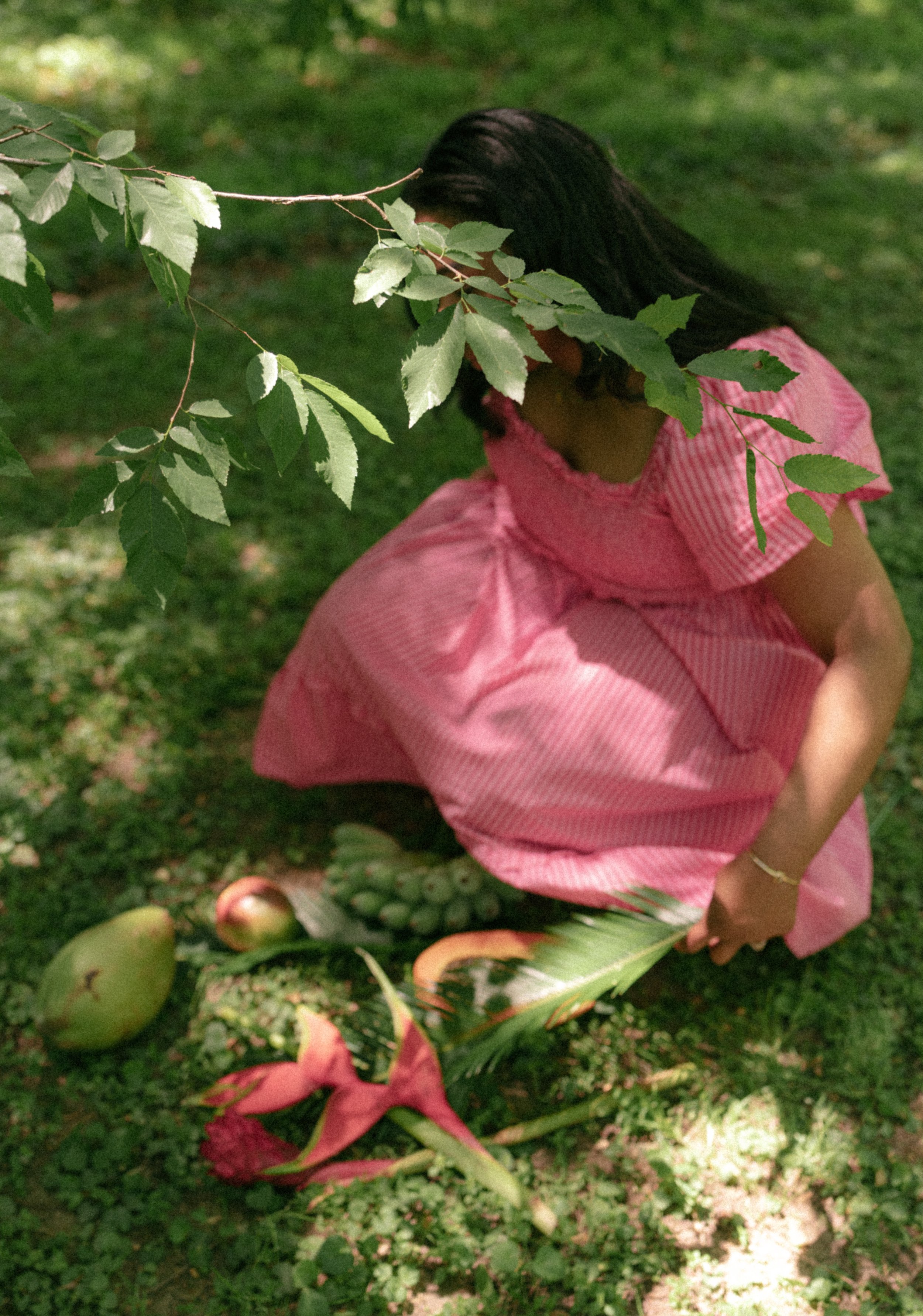 A young girl in a pink dress crouches behind a tree with green leaves, with apples on the ground nearby, in a park or garden setting.