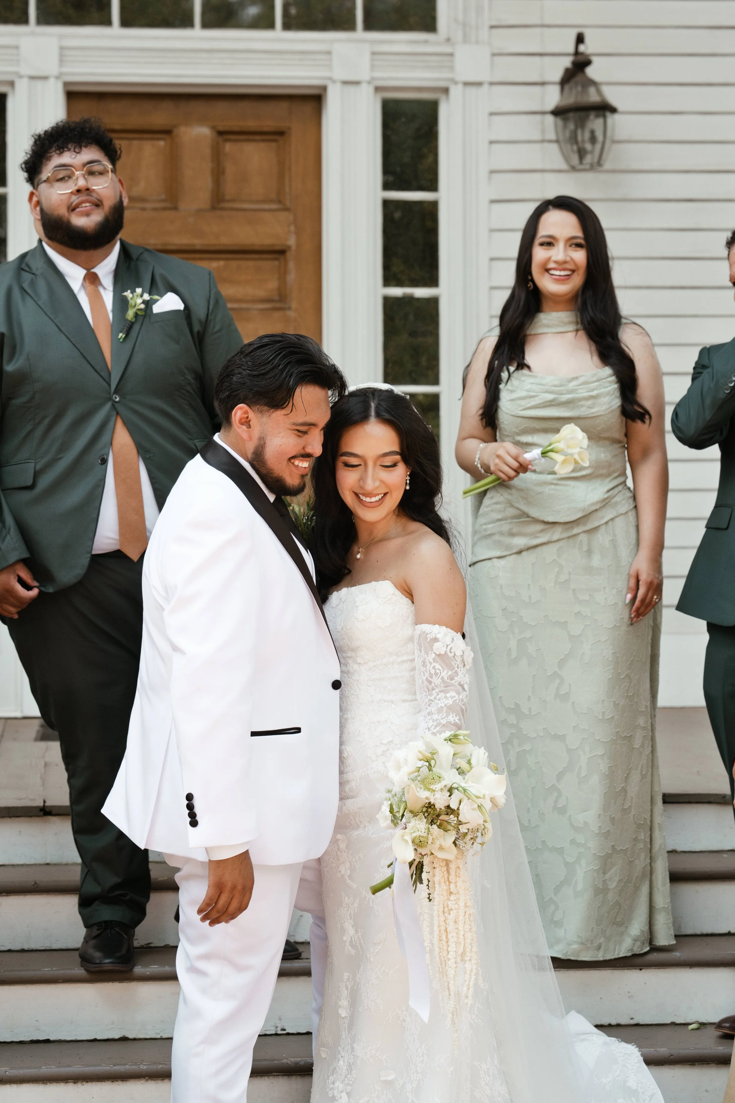 A wedding group photo on the steps of a house with three women and two men dressed in formal attire, smiling and holding flowers.