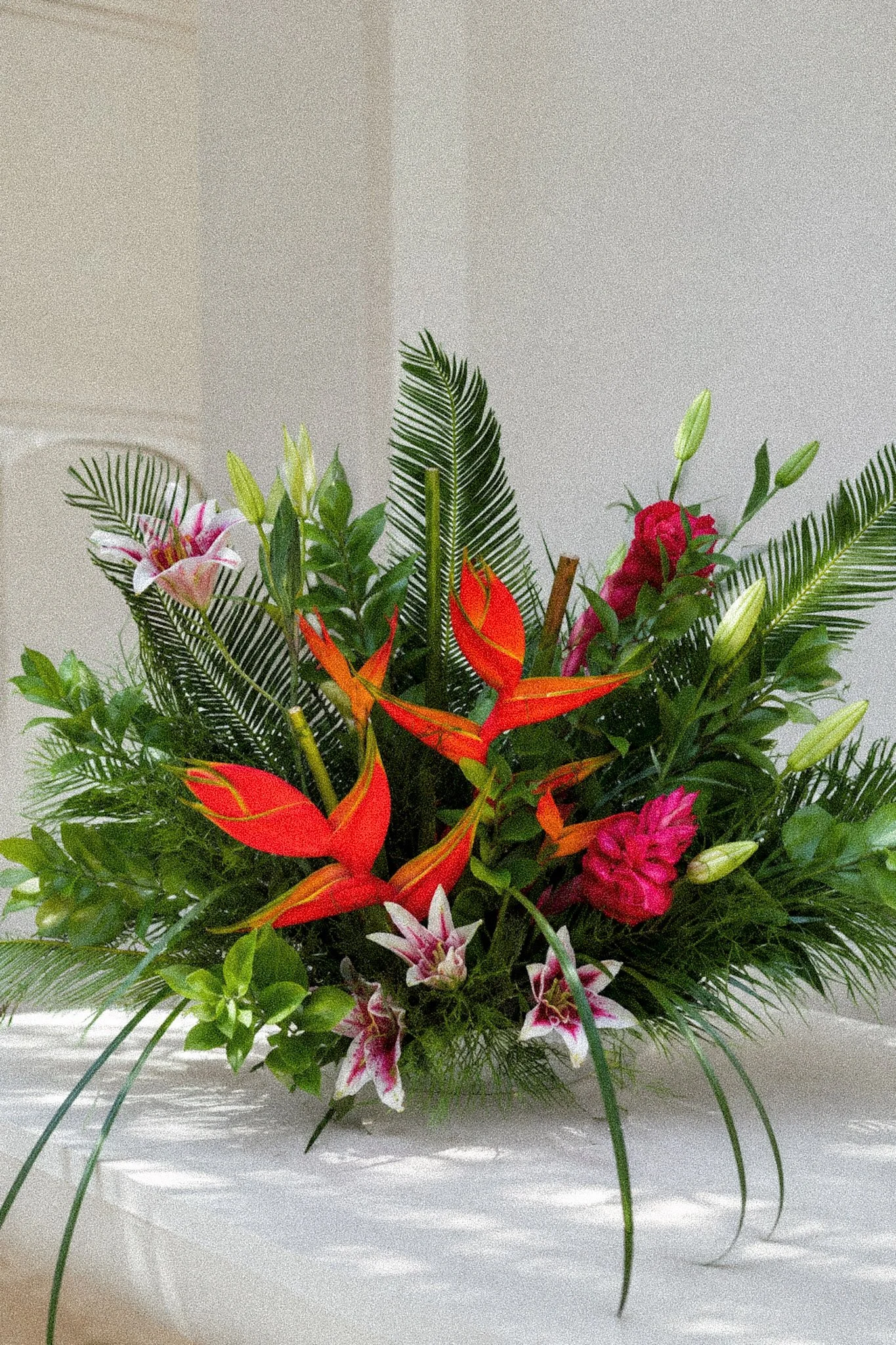 A colorful floral arrangement with pink lilies, red ginger flowers, white and pink lilies, green foliage, and palm leaves in a white vase.
