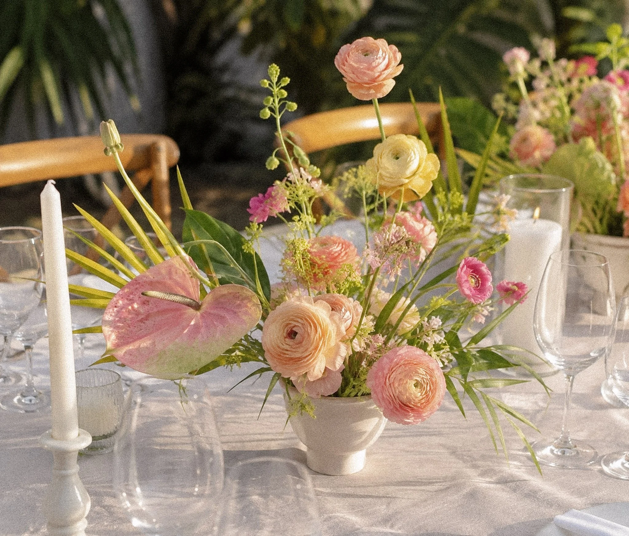 A floral table centerpiece with pink, yellow, and cream flowers in a white vase. Surrounding the arrangement are white candles and glassware on a table covered with a white tablecloth.