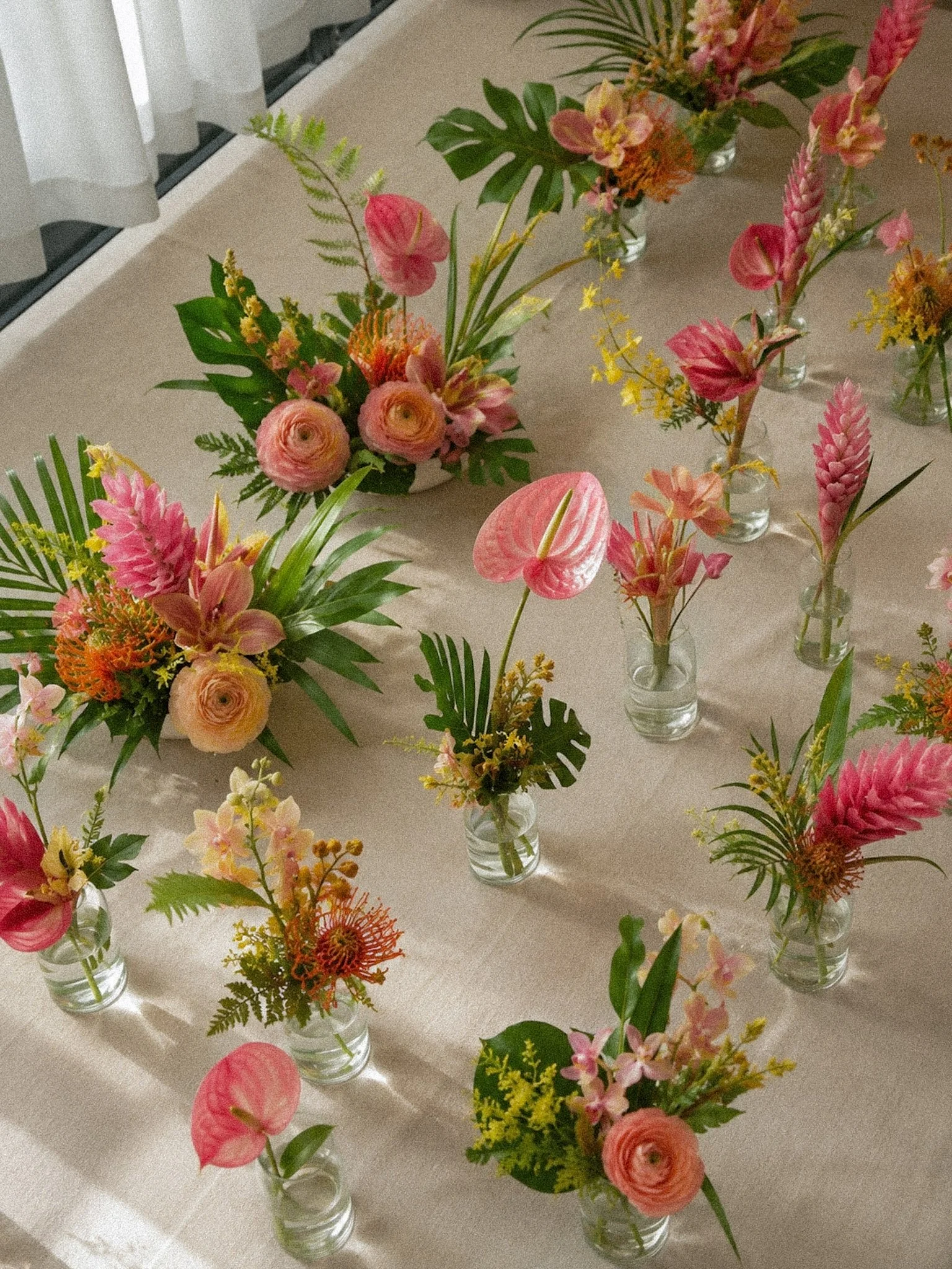 Multiple small vases containing colorful floral arrangements with pink, orange, and yellow flowers on a beige table, near a window with white curtains.