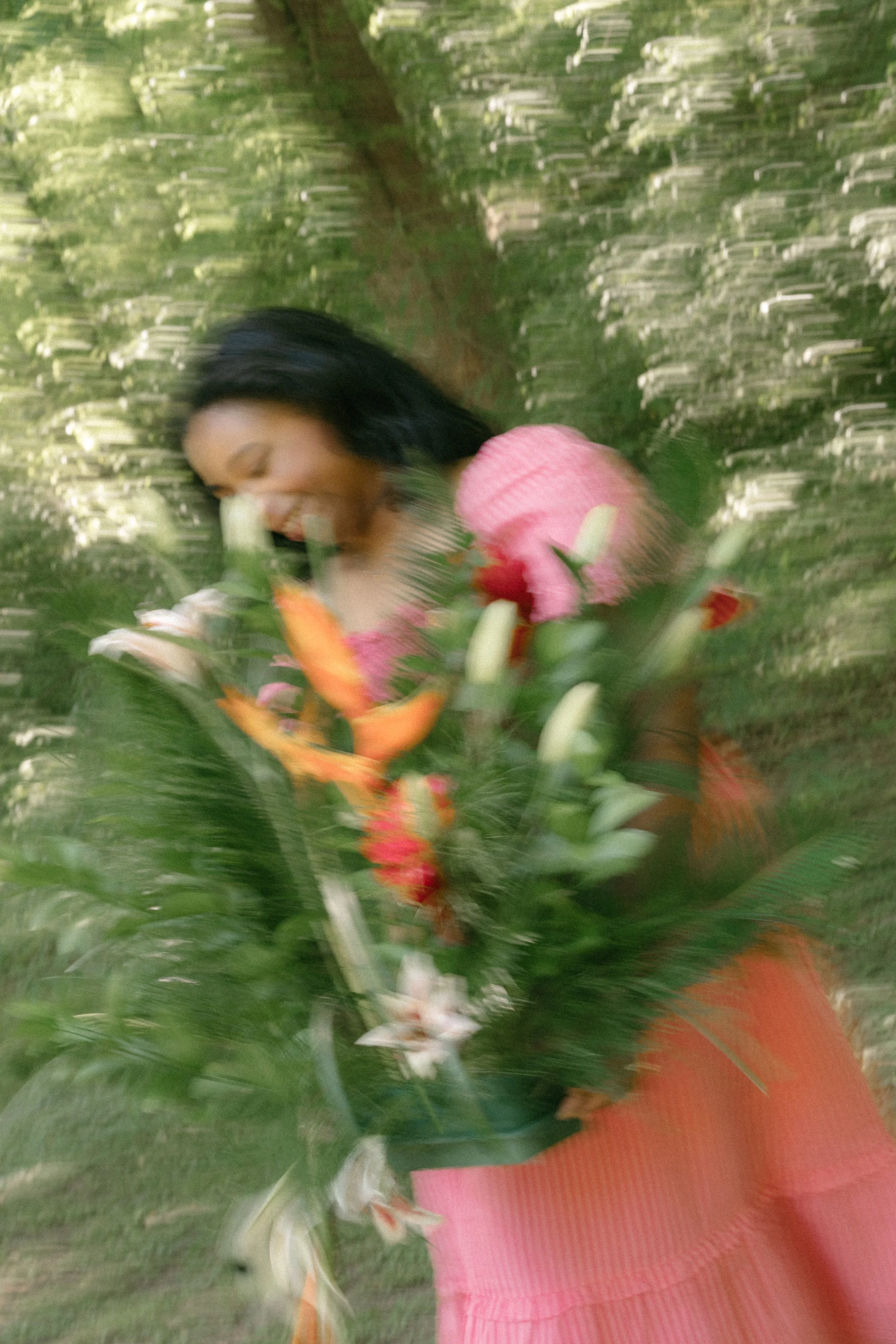 A woman with dark hair smiling and holding a bouquet of flowers, standing outdoors in a green, leafy environment.