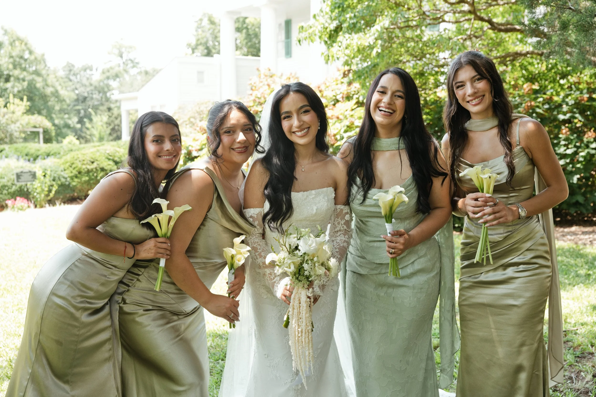 A bride and five women dressed in matching gold dresses, smiling and holding bouquets, standing outdoors on a sunny day with trees and a white house in the background.