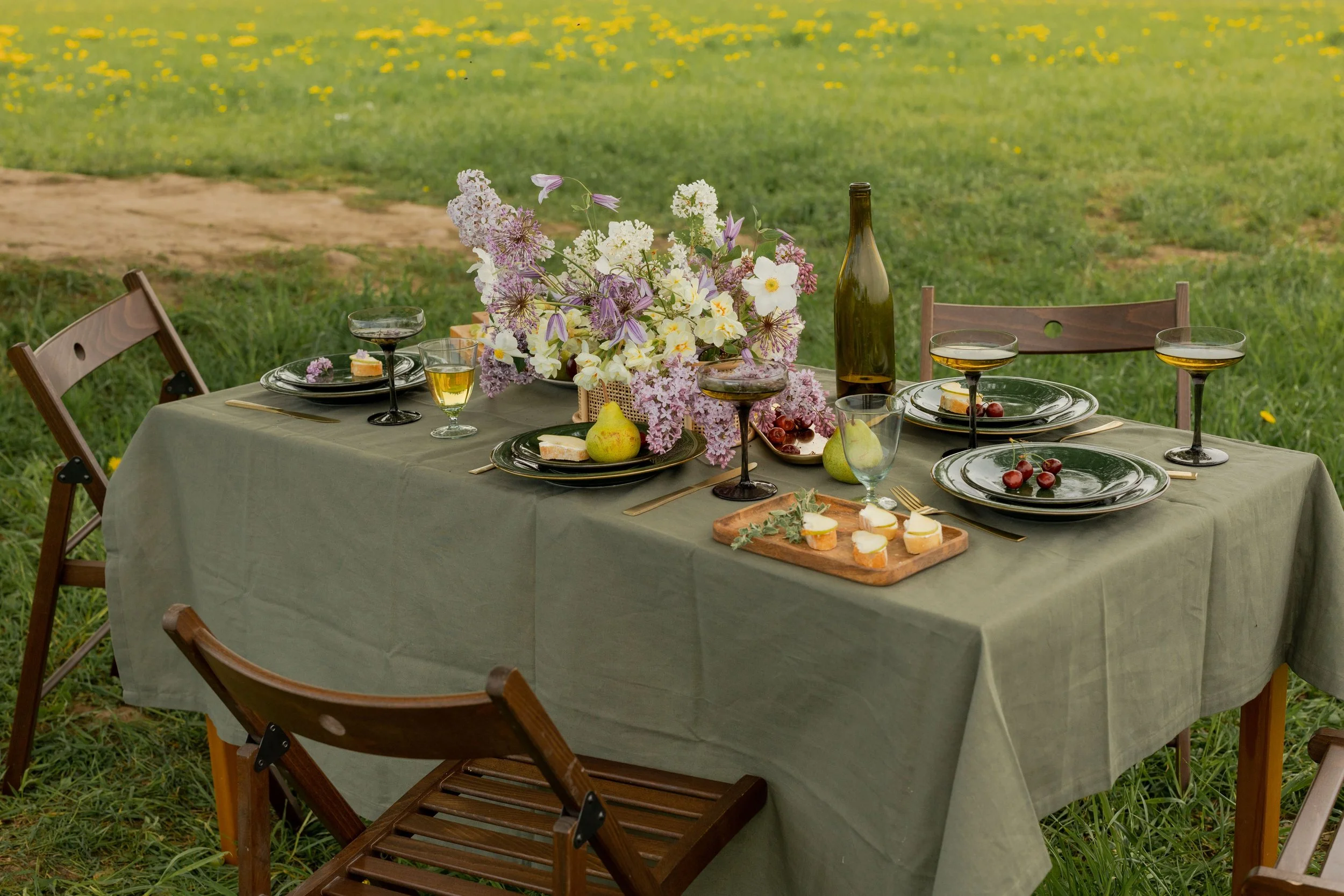 An outdoor table set with plates of cheese and grapes, a floral centerpiece, and glasses of white wine, on a green tablecloth, in a grassy field with yellow flowers.