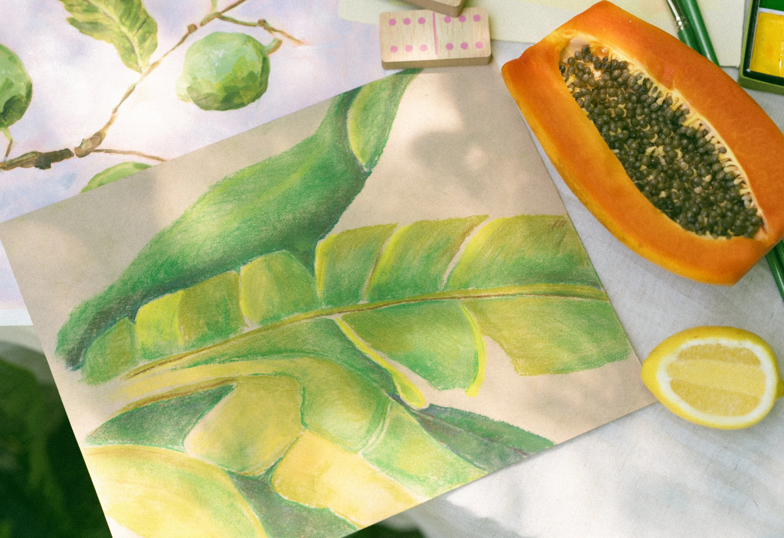 Close-up of a watercolor painting of green leaves and a branch, with art supplies including a halved papaya with black seeds, green pencils, a yellow lemon, and wooden stamping blocks nearby.