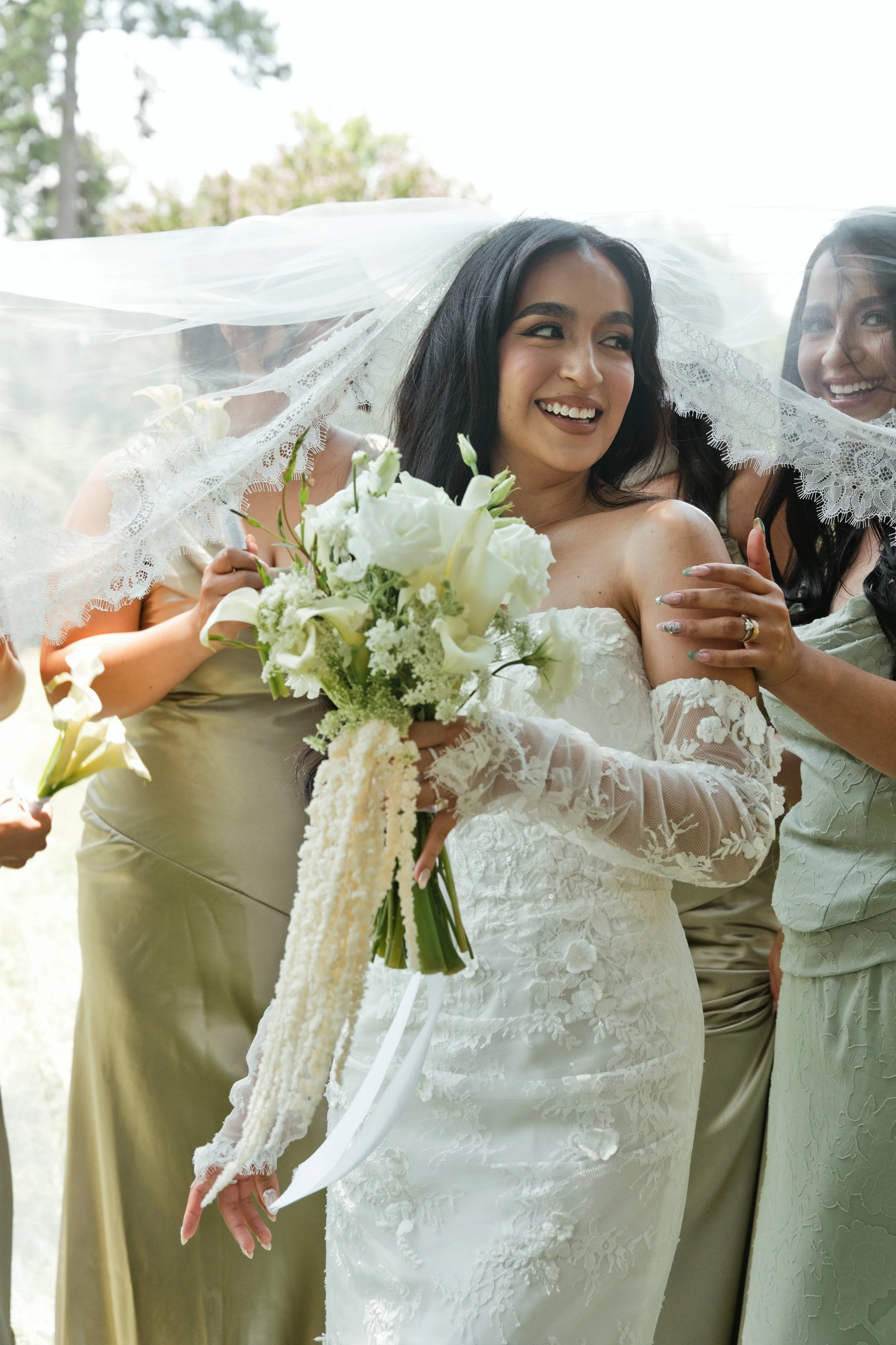 A bride in a white lace wedding dress, holding a bouquet of white flowers, smiling and leaning close to another woman under a lace veil with her friends during a wedding celebration.